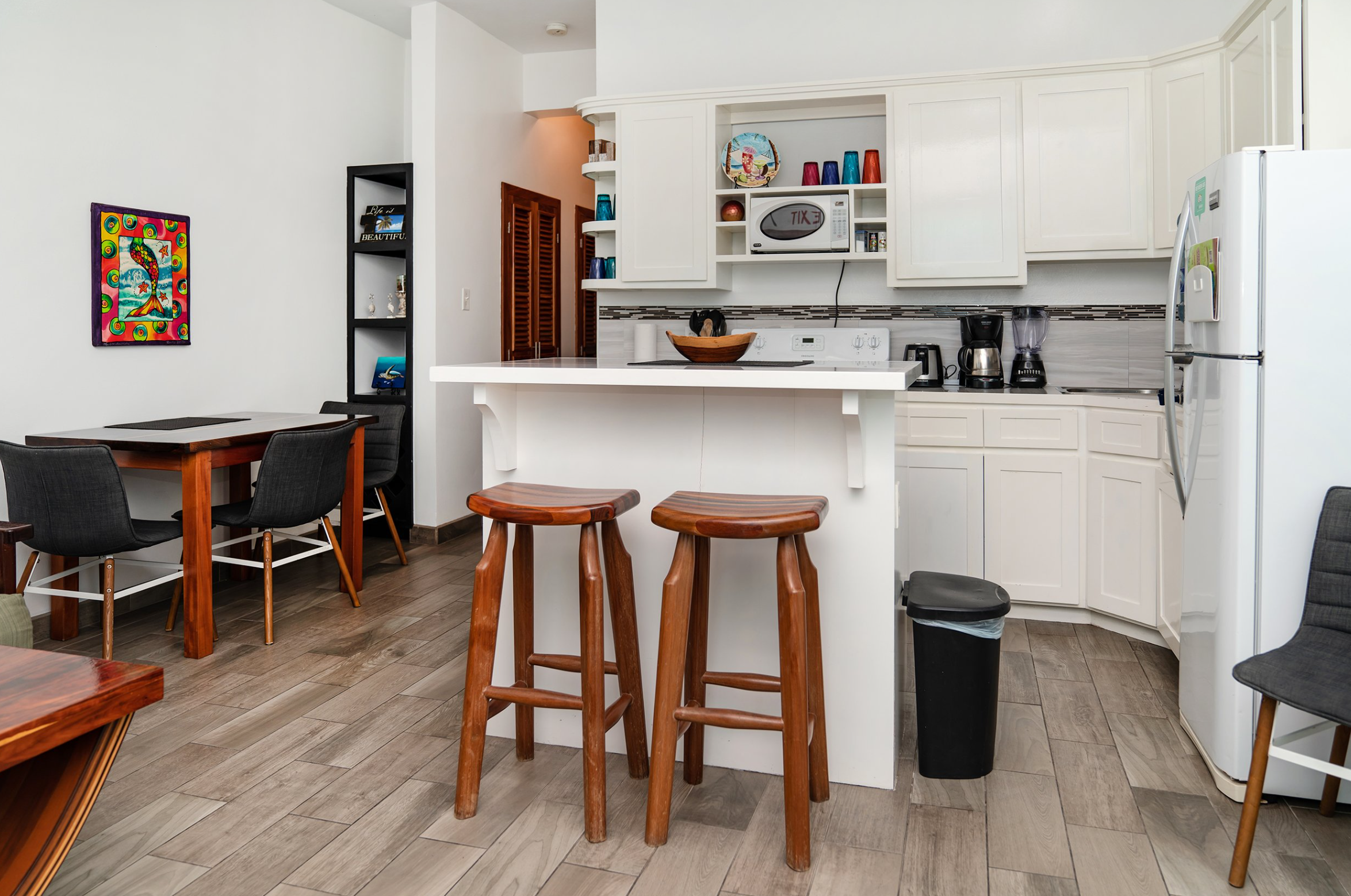 Modern kitchen and dining area with wooden stools at a white kitchen island, black chairs around a wooden dining table, and white cabinets. Colorful artwork and shelves with decor accents the space.