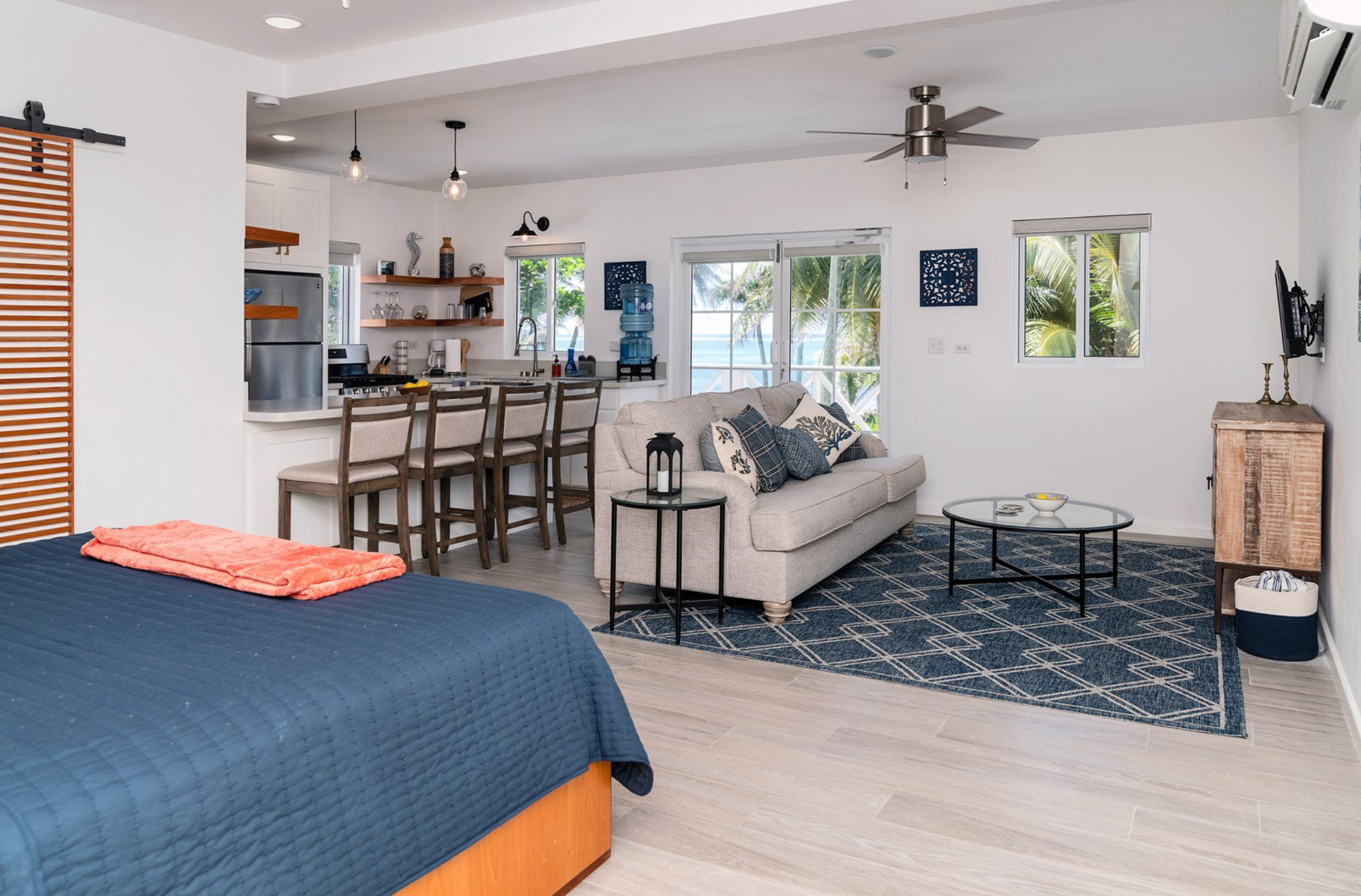 Modern open-concept living space with kitchen, dining area, and lounge. Features a bed with a blue quilt, a light gray sofa, a glass coffee table, and a view of palm trees and ocean through windows. Includes kitchen bar stools and pendant lighting.