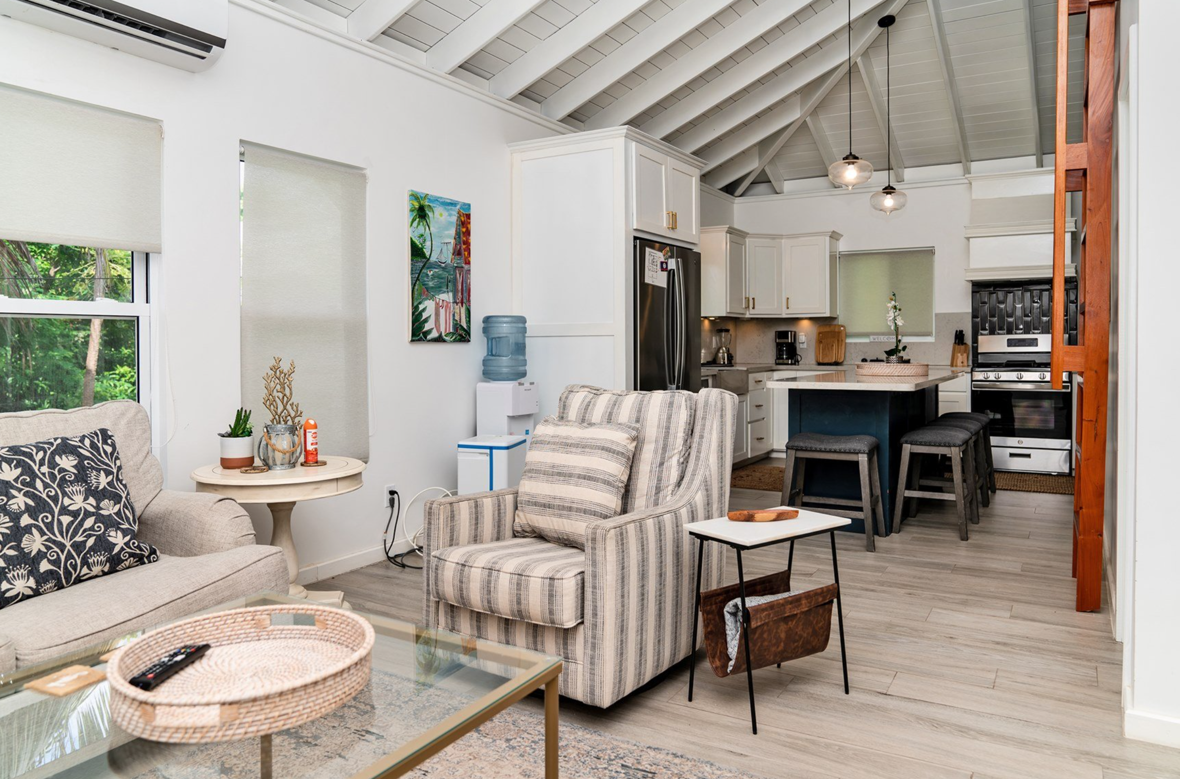 Cozy living room and kitchen with striped armchair, decorative pillows, plants on side table, modern kitchen island with barstools, stainless steel refrigerator, and decorative wall art.