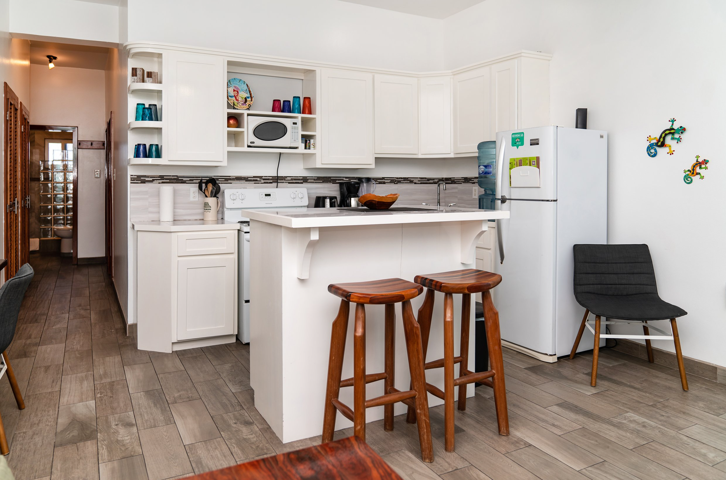 Modern kitchen with white cabinets, bar counter, wooden stools, refrigerator, microwave, and colorful decorations.