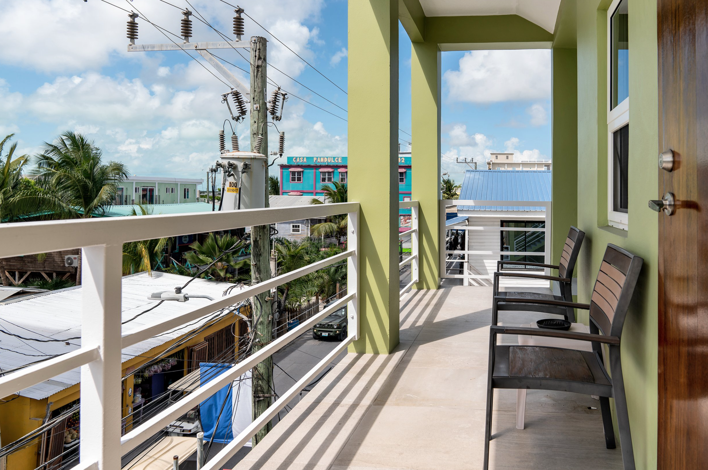 View from a sunny balcony with green walls, metal railings, and two chairs. Overlooking a street with buildings, palm trees, and power lines. Cloudy blue sky in the background.