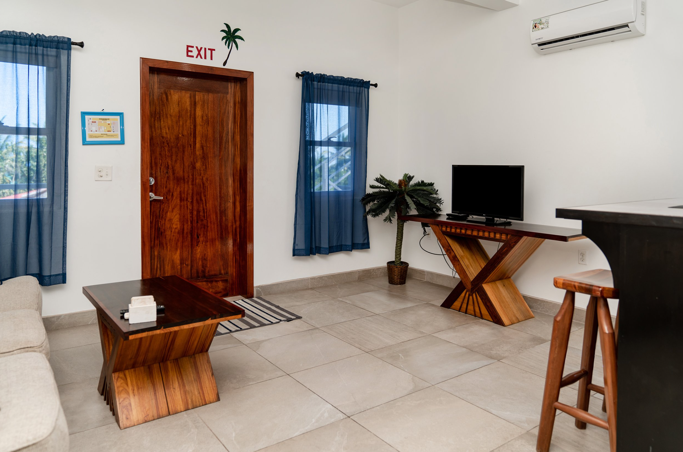 Living room with wooden furniture, including a TV on a console, a coffee table, and a bar stool. Two windows with blue curtains, a potted plant, and a split air conditioner mounted on the wall. San Pedro, Boca Del Rio location