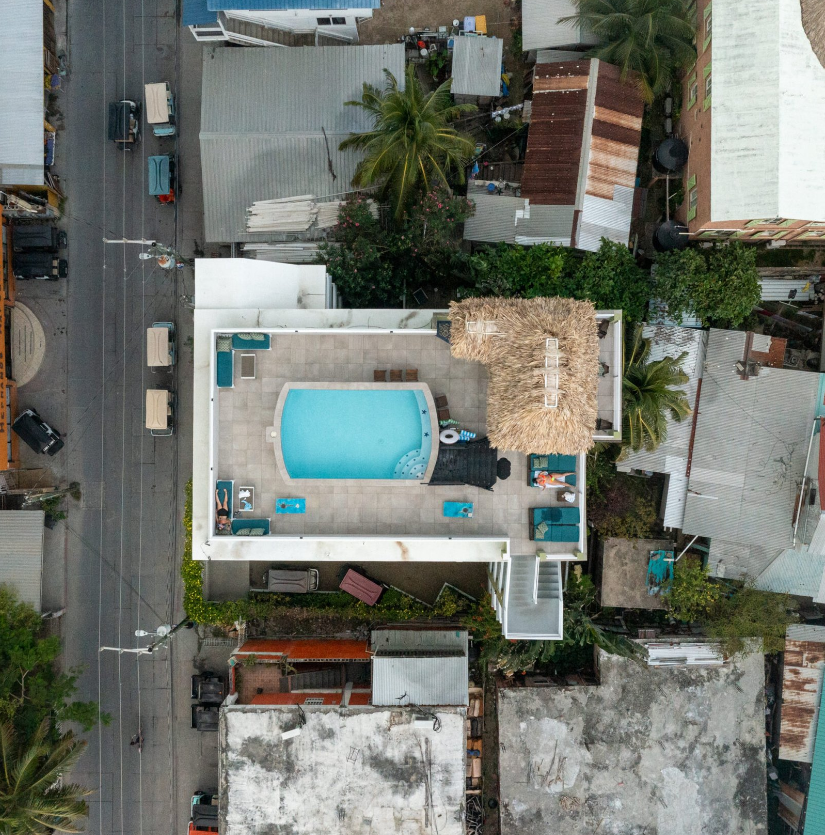 Aerial view of a rooftop with a swimming pool, lounge chairs, and a palapa in an urban neighborhood. Surrounding buildings and a street are visible.