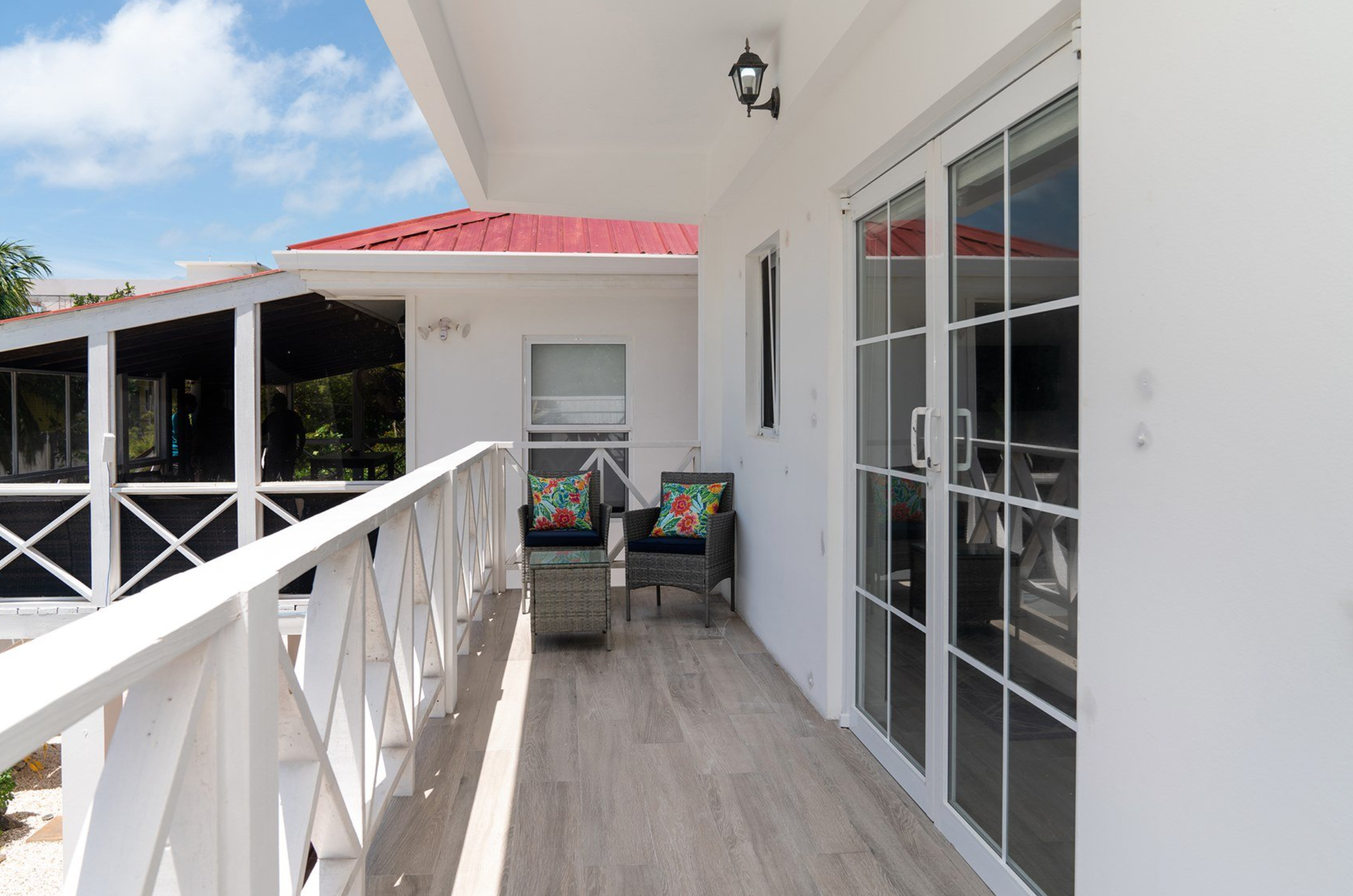 Balcony with wicker chairs and colorful cushions, glass sliding doors, and white railing. Red roof and blue sky in the background.