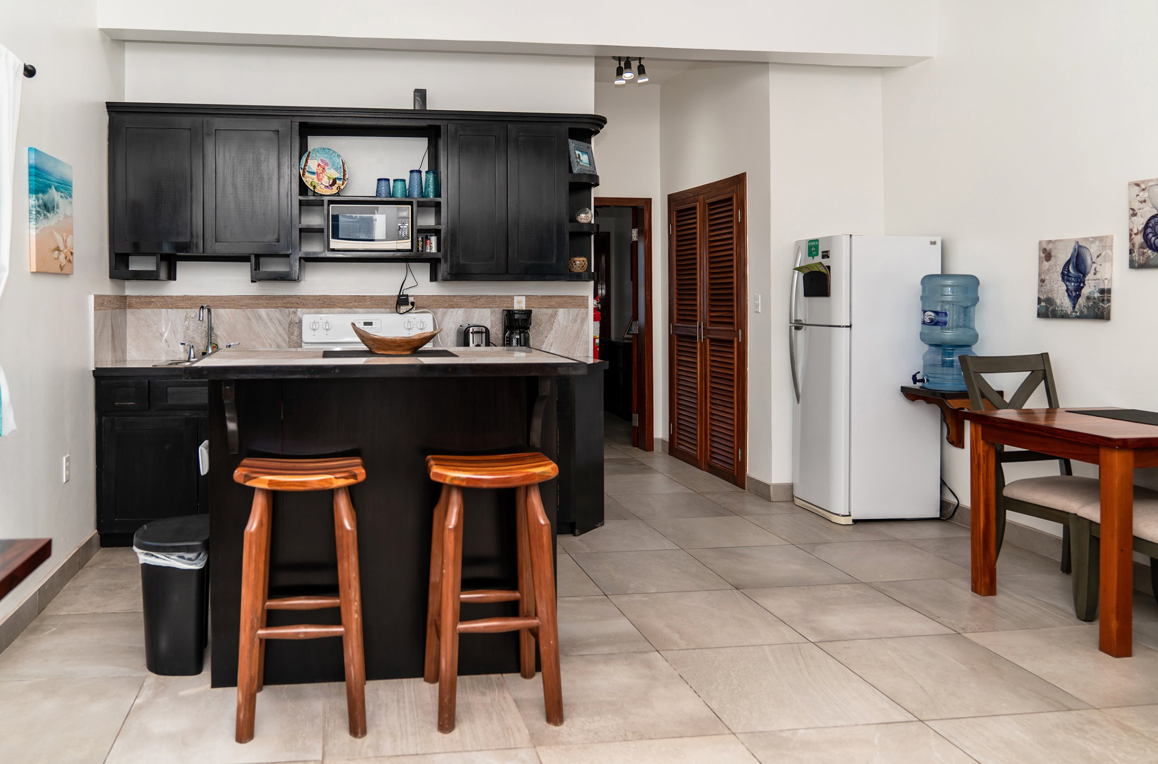 Modern kitchen with black cabinets, wooden bar stools, refrigerator, water dispenser, dining table, and wall art.