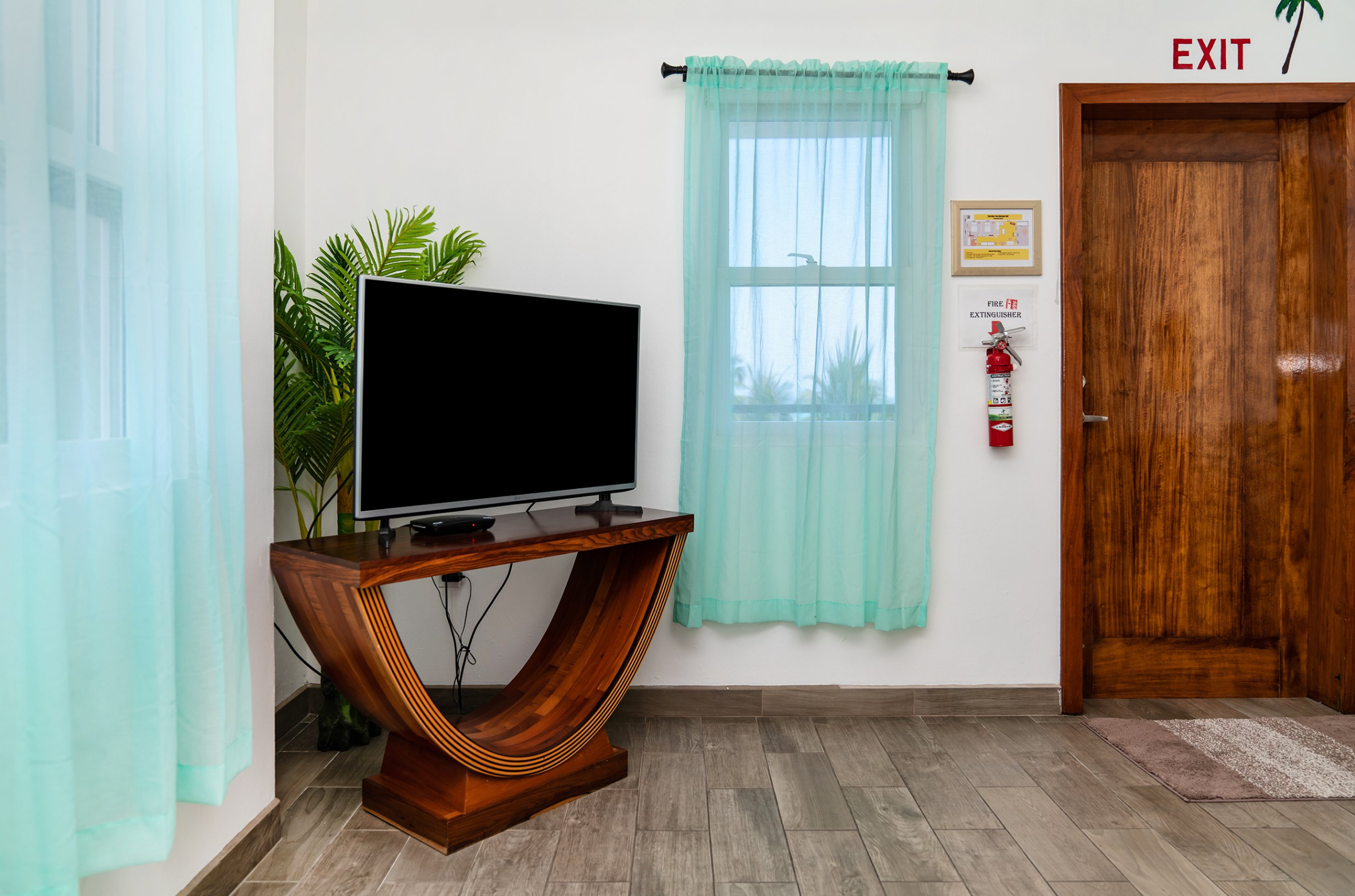 Room corner with a TV on a curved wooden stand, green sheer curtains, a fire extinguisher on the wall, and a wooden door with an exit sign above.