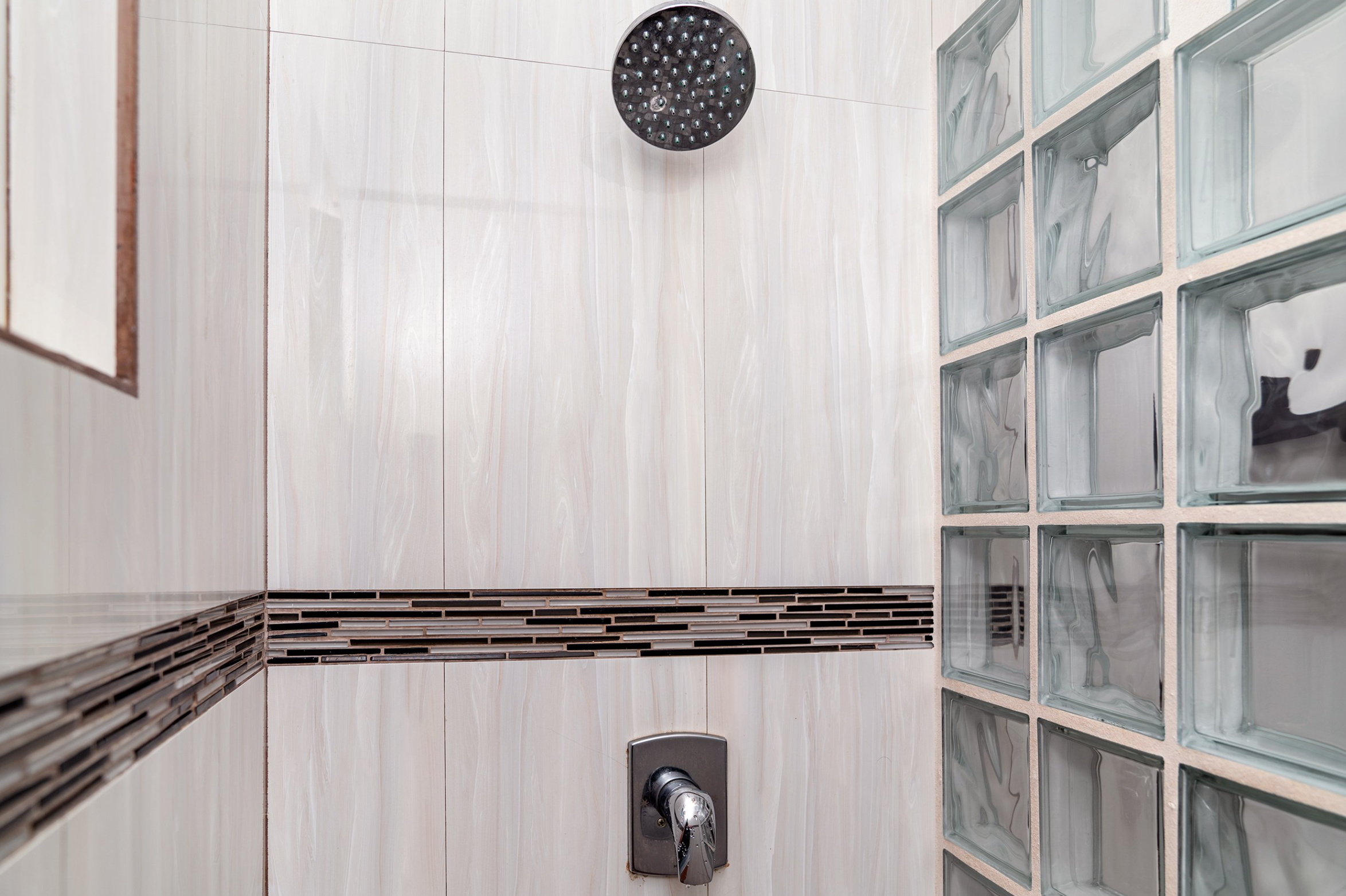 Modern bathroom shower with wall-mounted showerhead, large beige tiles, decorative horizontal tile accent, and glass block divider.