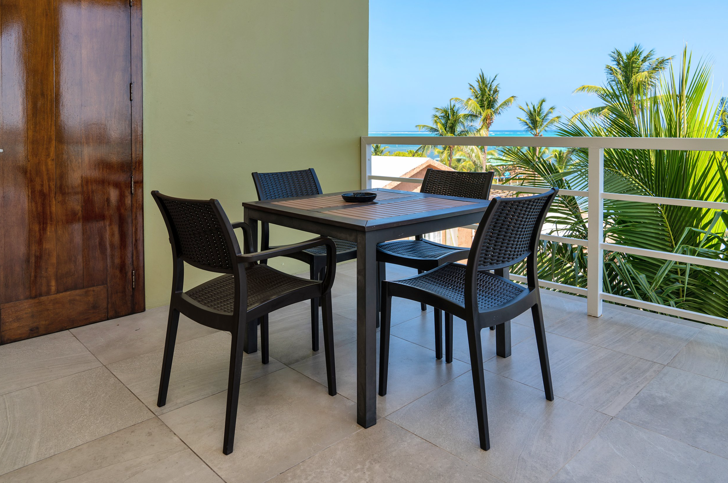 Outdoor patio with a square table and four black chairs, with palm trees and ocean view in the background.