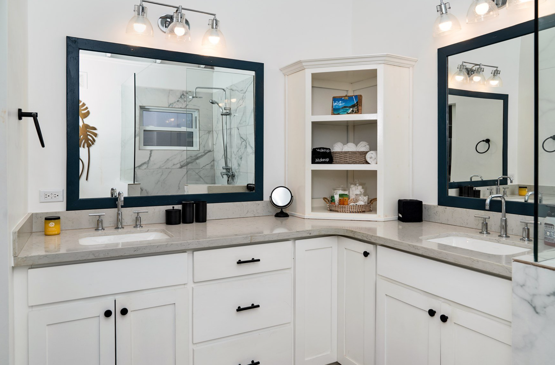 Modern bathroom vanity area with two sinks, marble countertops, large mirrors, and open shelving. The shelves contain towels and toiletries. Overhead lighting and a shower reflected in the mirrors.