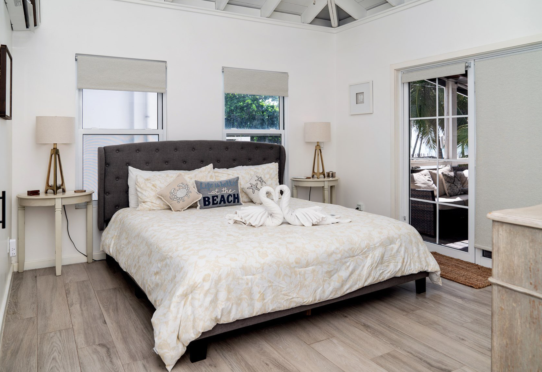 Coastal-themed bedroom with a large bed, decorative pillows, towel swans, bedside tables, and lamps. Sliding glass doors open to a patio area with tropical views.