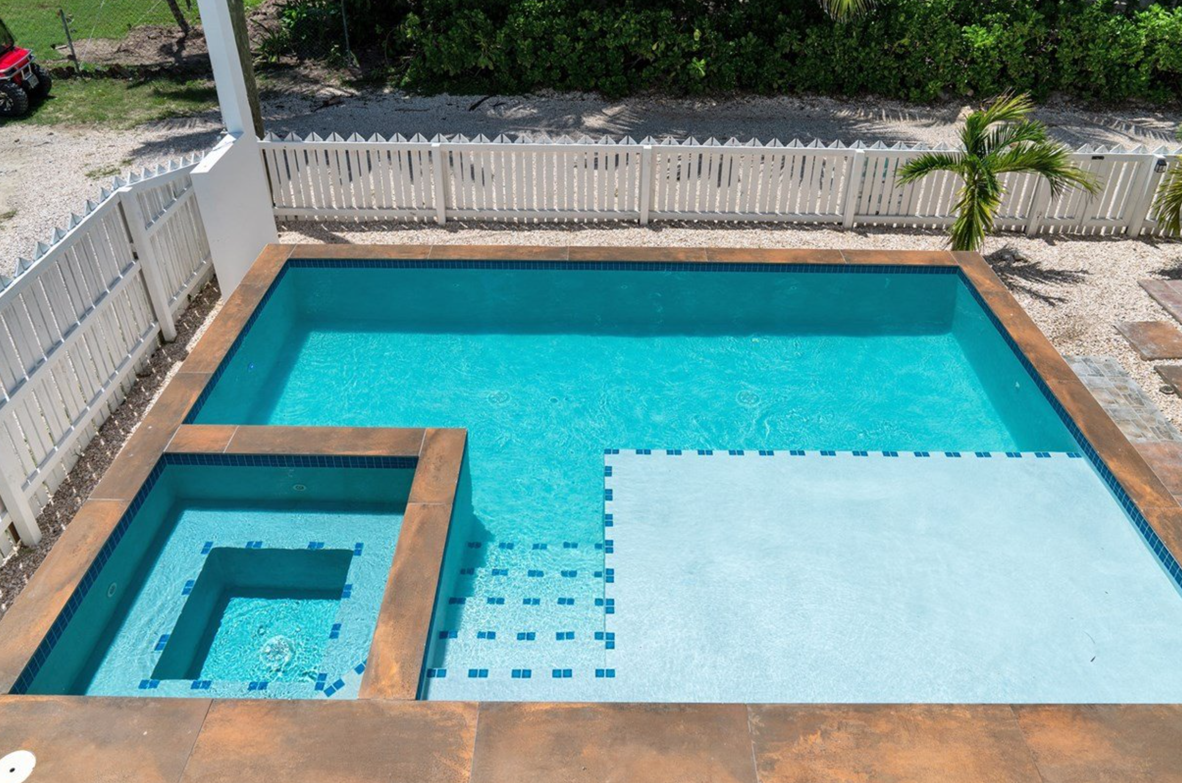 Overhead view of a rectangular swimming pool with an attached hot tub, surrounded by a white picket fence and greenery.