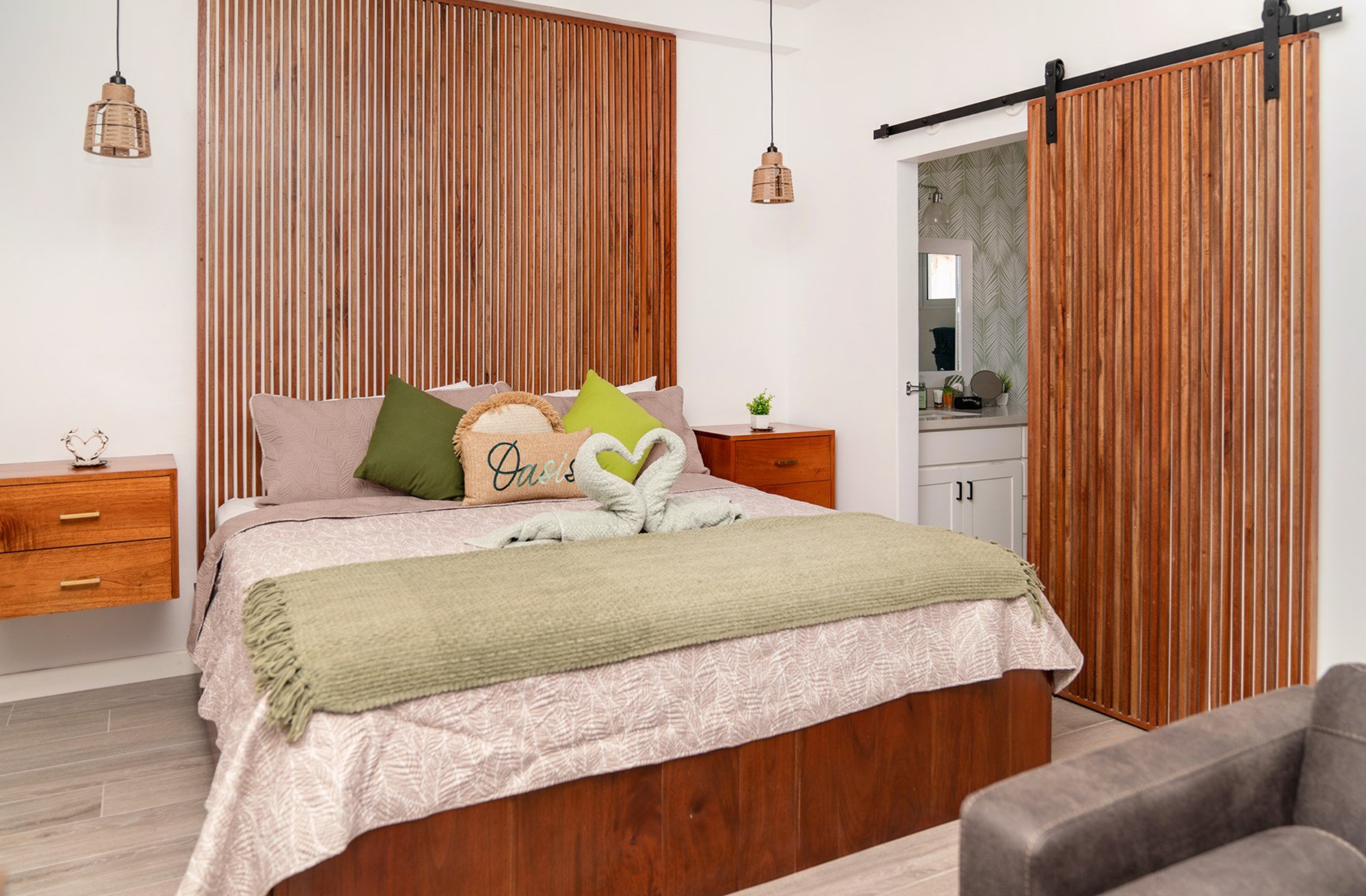 Modern bedroom with wooden accents, featuring a bed with green and beige pillows, a green throw, a wooden slat headboard, hanging pendant lights, and a sliding wooden door leading to a bathroom.