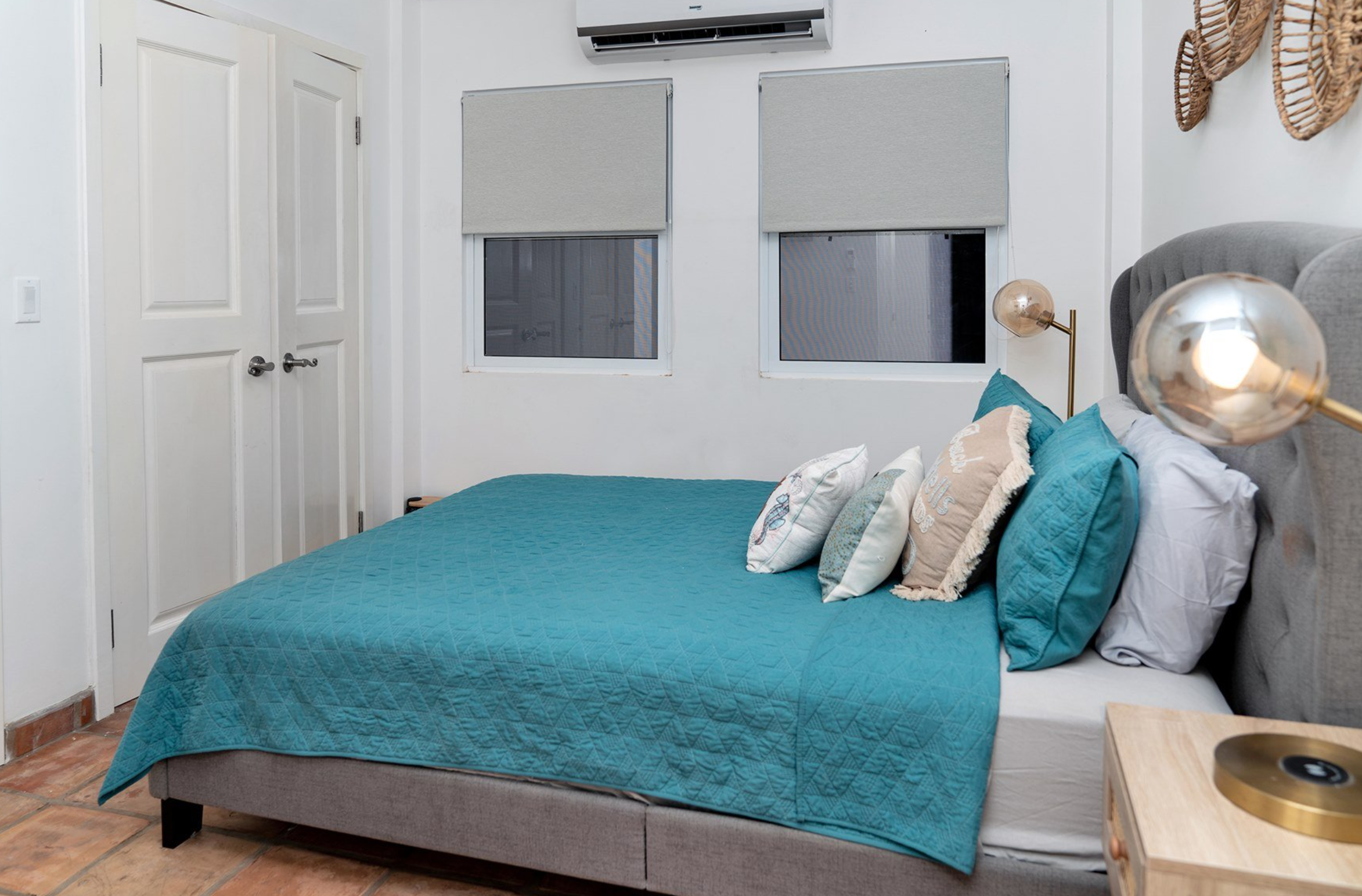 Bedroom with teal bedding, gray headboard, and decorative pillows on the bed. Two windows with shades and wall-mounted air conditioner above. Bedside table with brass lamp and woven wall decor.