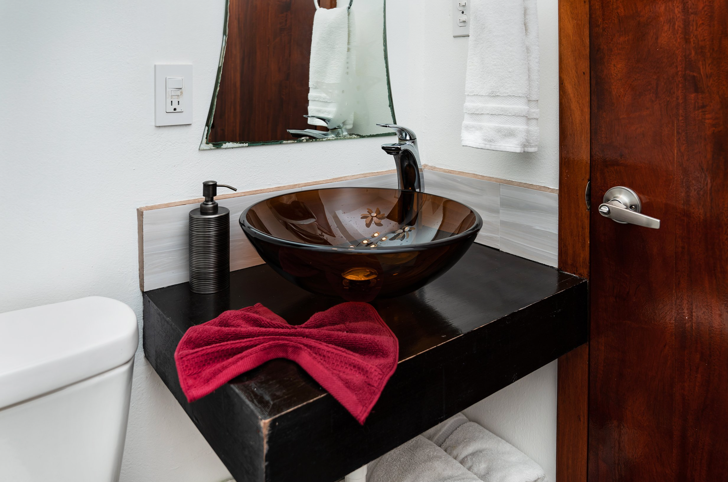 Modern bathroom sink with brown glass vessel basin, chrome faucet, black soap dispenser, red towel on counter, and mirror above. Towel rack and dark wooden door visible in the background.