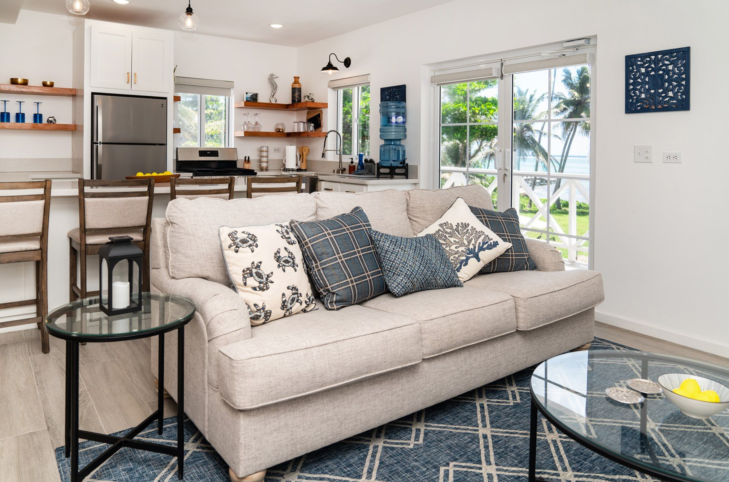 Coastal themed living room with gray sofa and blue patterned cushions, glass-top tables, and a view of palm trees through large windows. Kitchen with silver appliances in background.