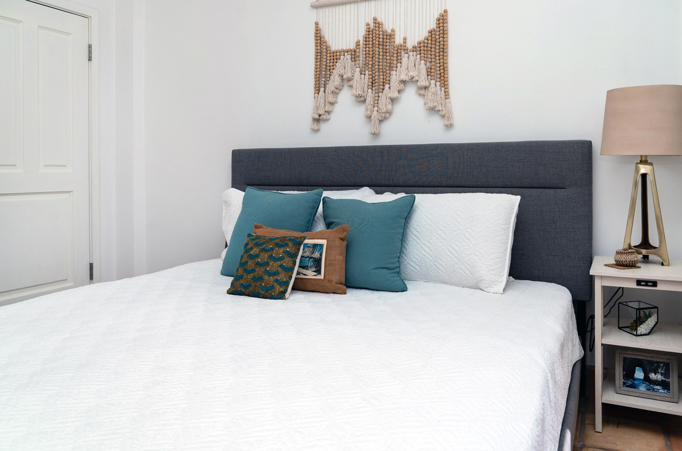Modern bedroom with blue and white bedding, decorative pillows, a gray upholstered headboard, a macrame wall hanging, and a nightstand with a lamp and decor items.