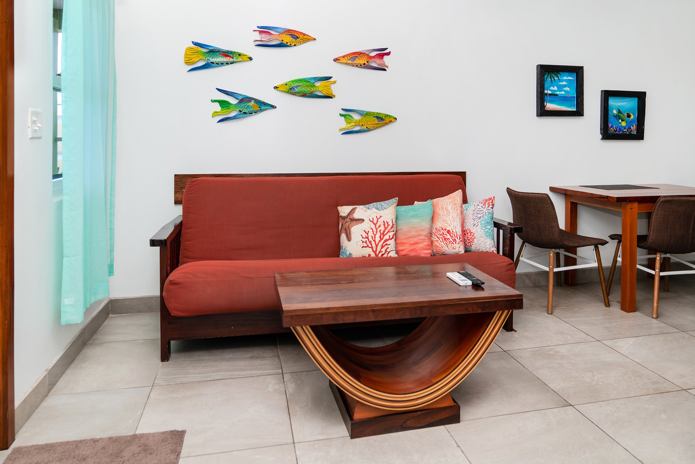 Living room with red sofa, colorful pillows, wooden coffee table, wall-mounted fish decorations, and a small dining table with two chairs.