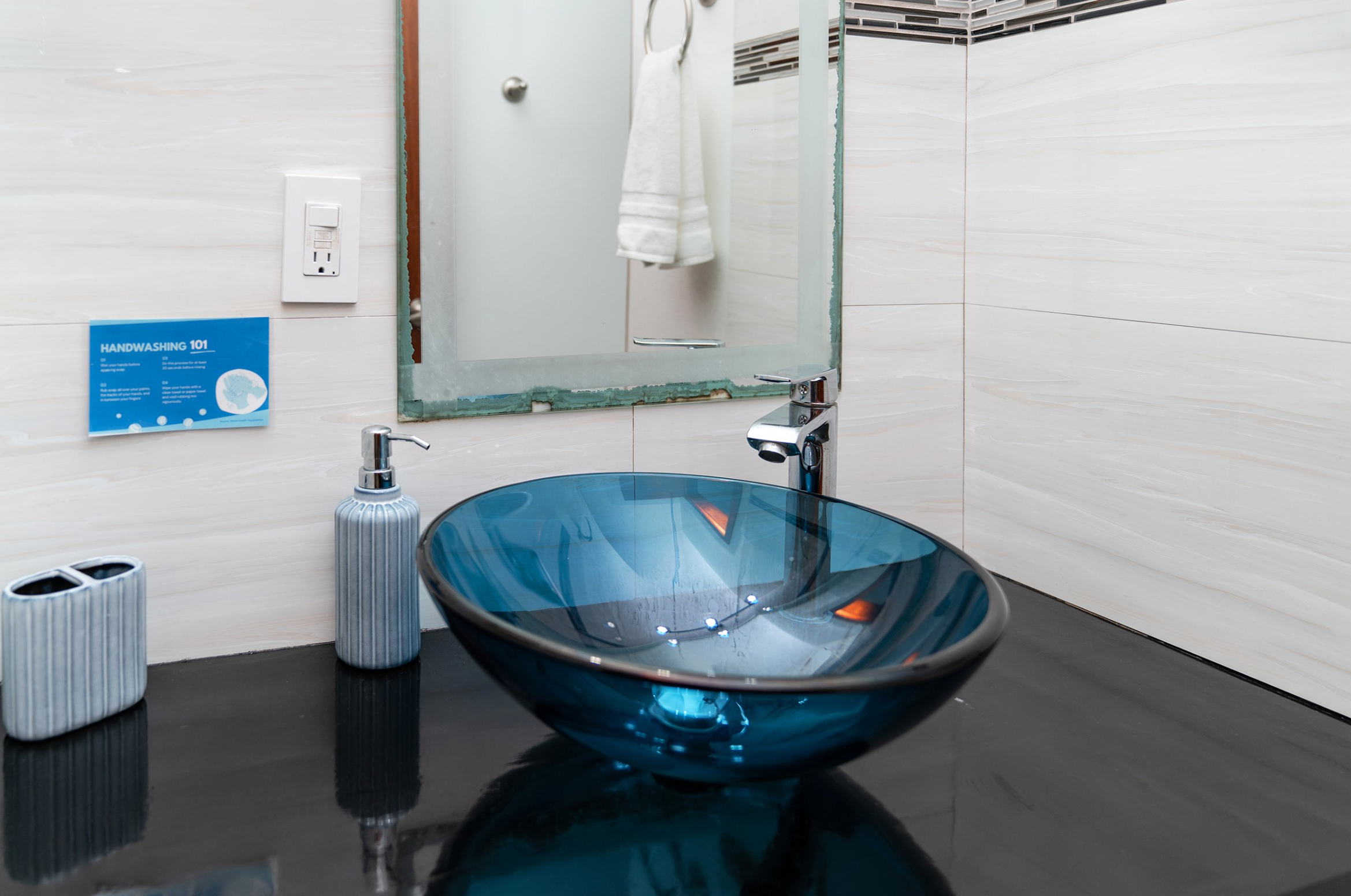 Bathroom countertop with a blue glass sink, soap dispenser, toothbrush holder, mirror, and a handwashing sign.