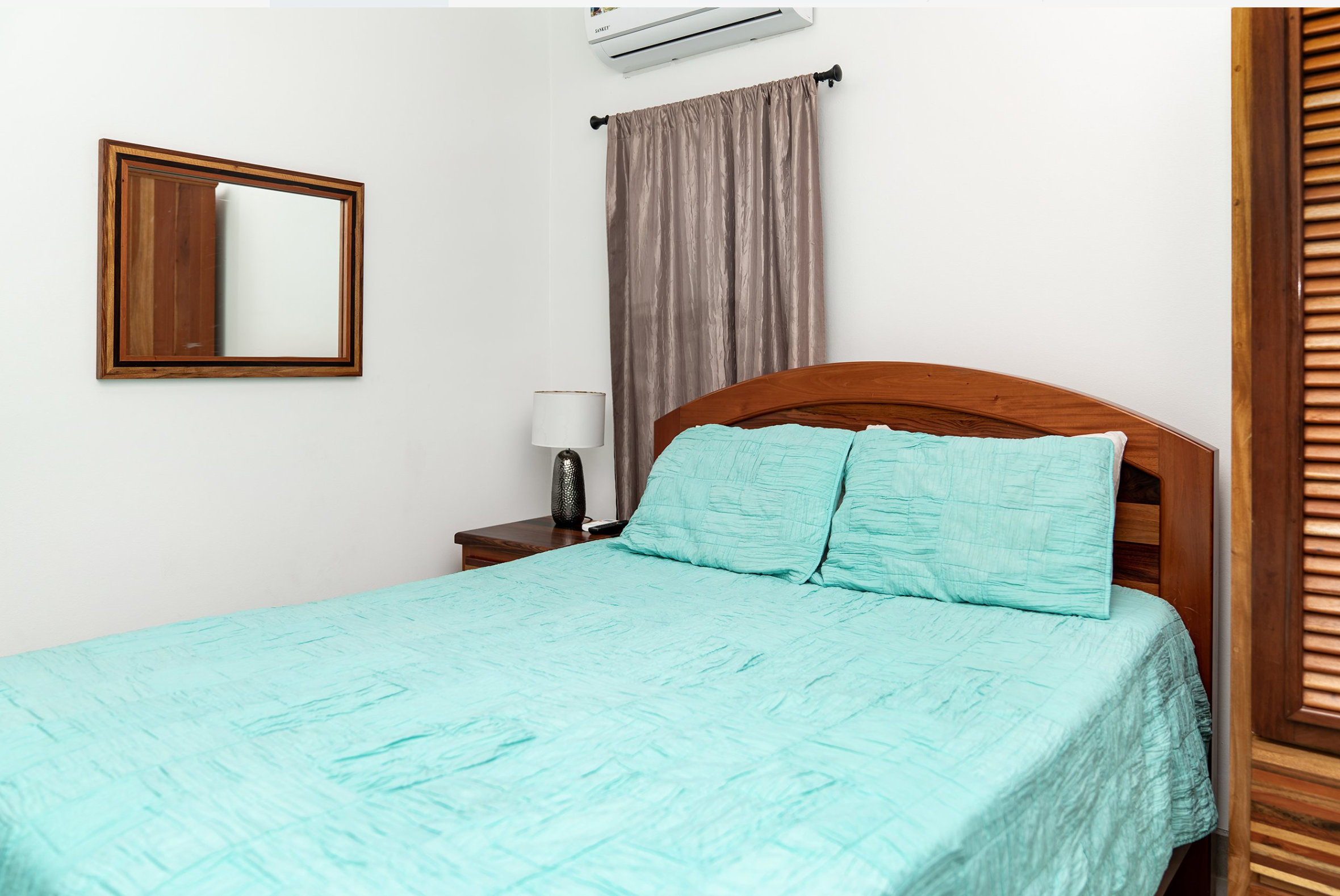 Bedroom interior with a wooden bed frame and a mint green quilt. A bedside table with a lamp is on the left, and a rectangular mirror hangs on the wall. A curtain covers the window above the bed.