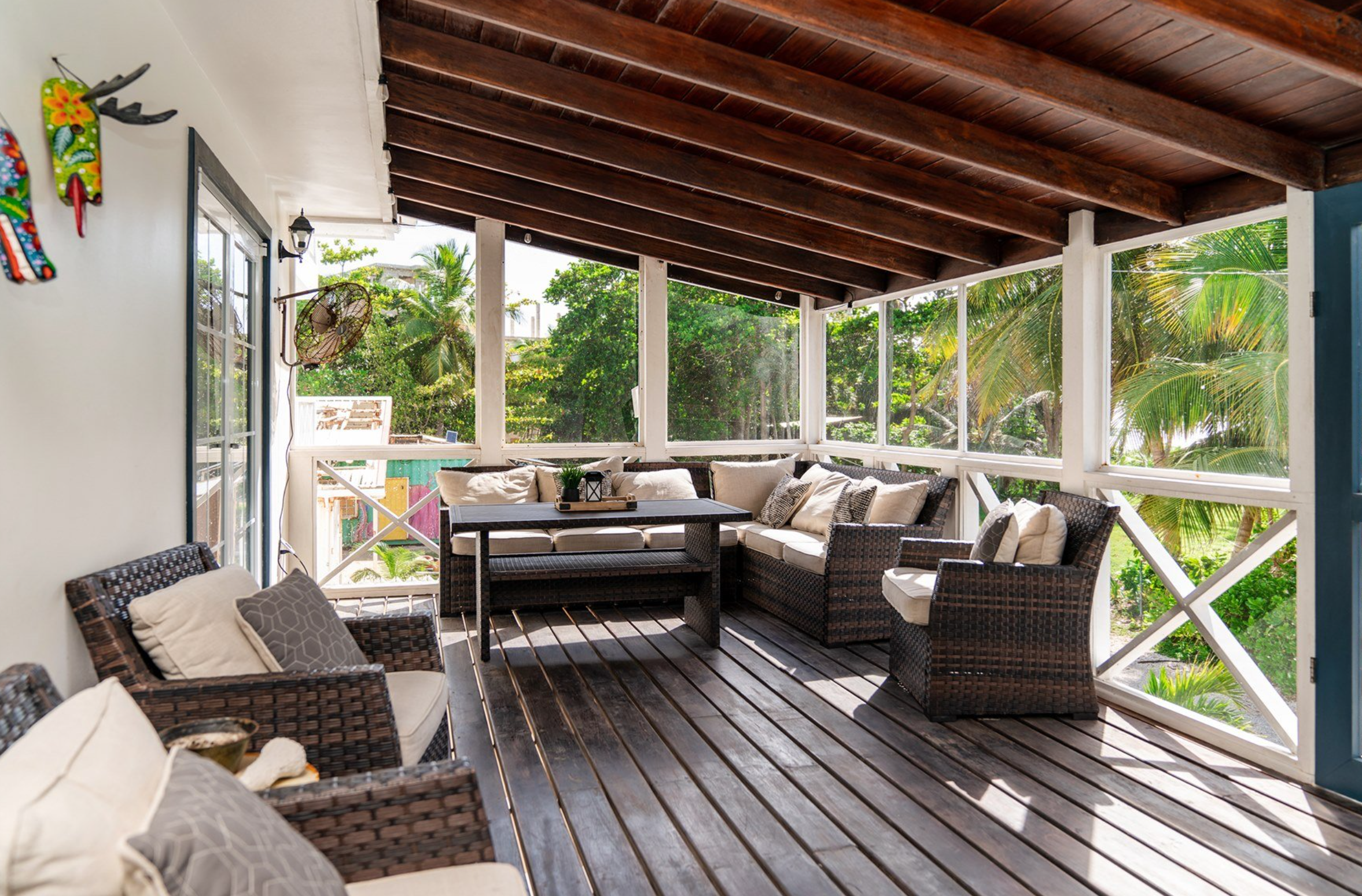 Covered patio with wicker furniture, pillows, and a small table, overlooking a lush garden, wooden roof, and decorative wall art.
