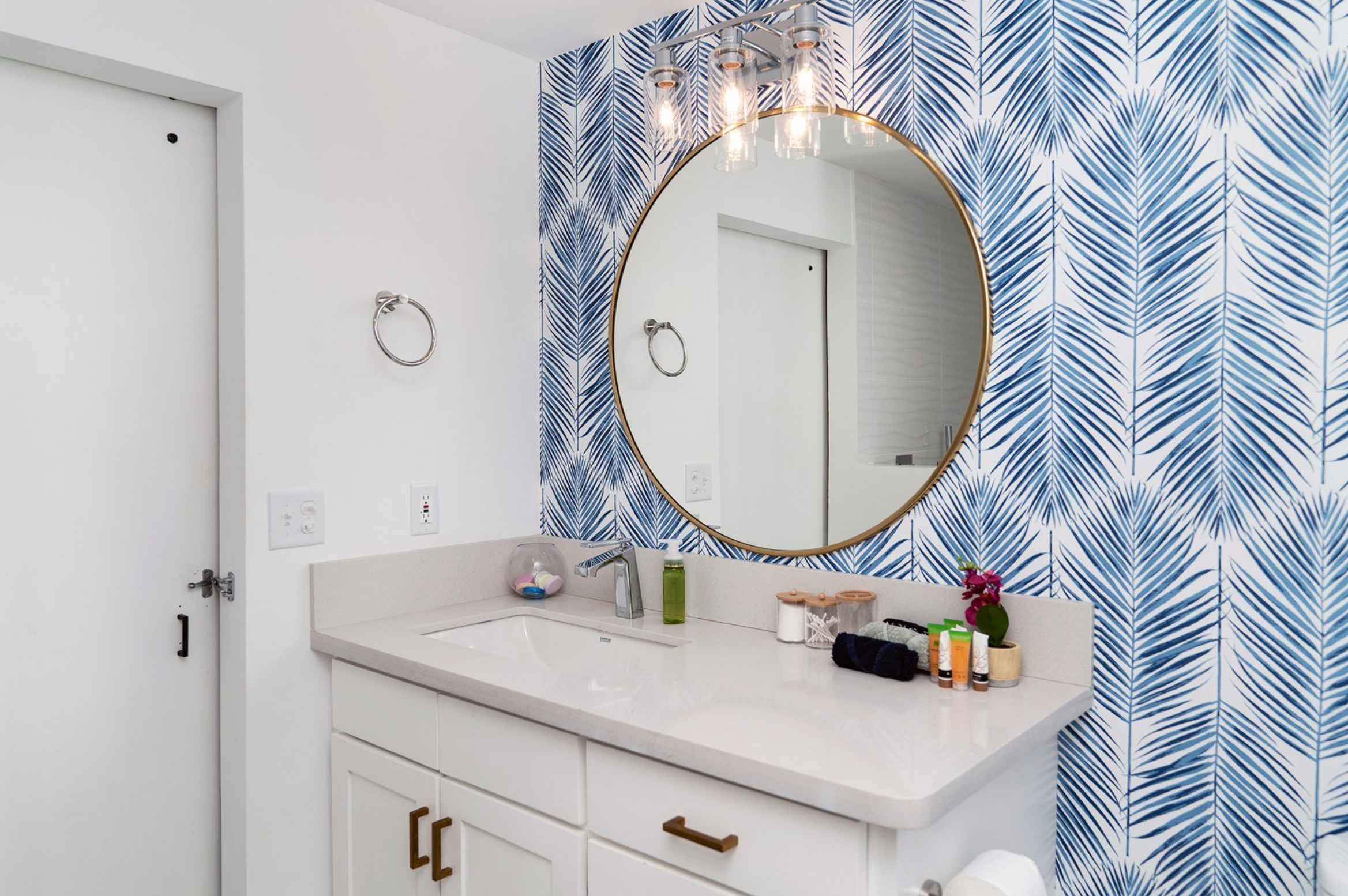 Modern bathroom with blue palm leaf wallpaper, round mirror, vanity with white countertop, and various toiletries.