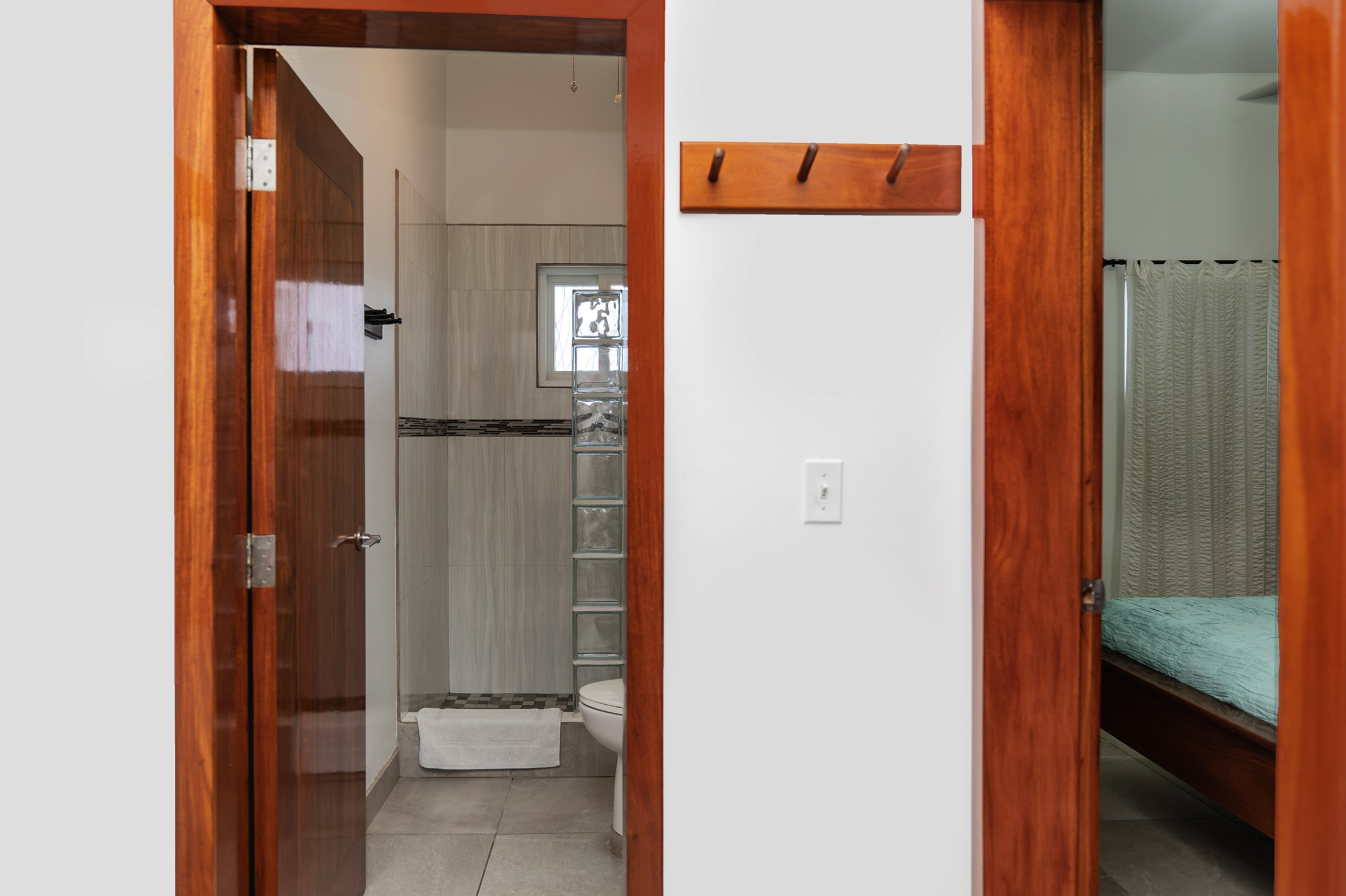 Interior view showing a bathroom and bedroom. The bathroom has tiled walls and floor with a small window and a towel. The bedroom has a wooden bed with a blue cover. Both rooms have wooden doors, and there is a coat rack and light switch on the wall in between.