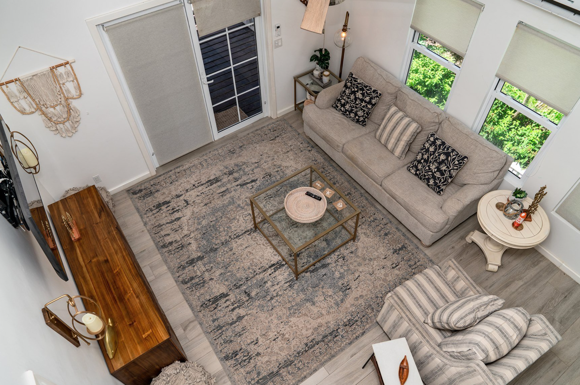 Modern living room with gray sofa, patterned pillows, glass coffee table, and decorative rug, viewed from above. Wooden console table and wall decor on the left, armchair at the bottom.