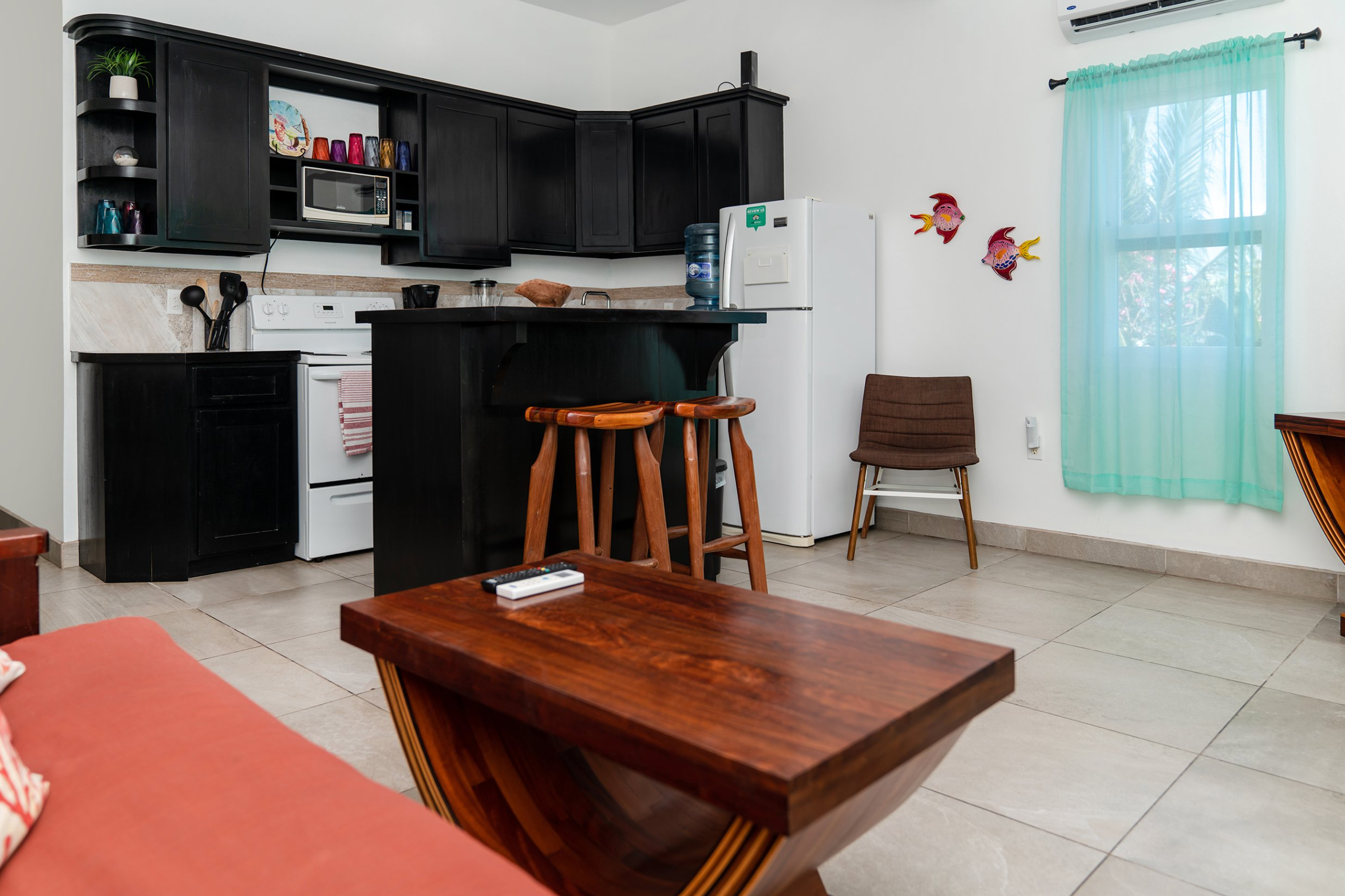 Modern kitchen with black cabinets, white stove, and refrigerator. Wooden stools are at the counter, and a small chair sits by a window with teal curtains. Colorful fish decorations are on the wall Excellent location, very walkable to the san pedro