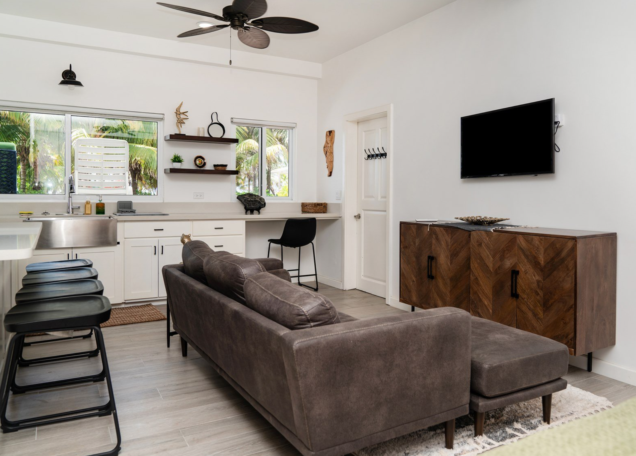 Modern living room and kitchen with a brown sofa, white cabinets, wall-mounted TV, and ceiling fan. Ambergris Caye property for sale.