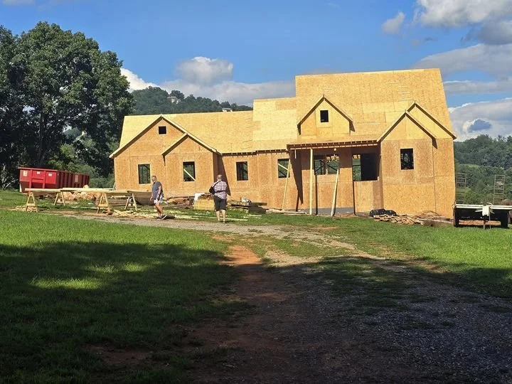 A house under construction with wooden framing, with a clear sky and trees in the background, and workers on site.