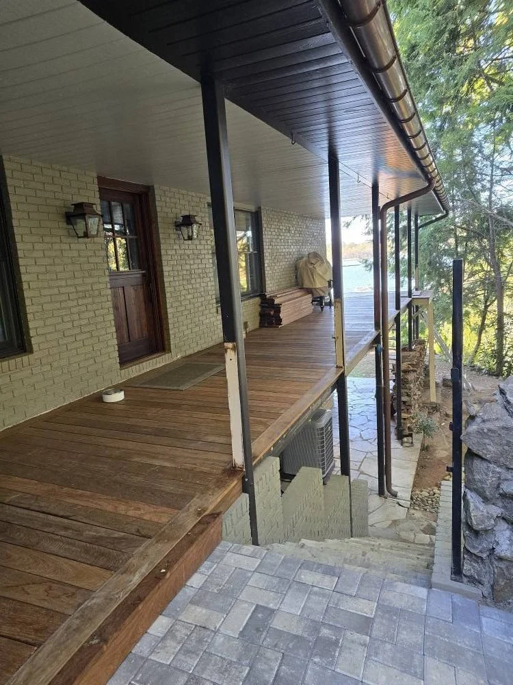 Wooden porch with stairs leading down, attached to a house with a brick wall, window, and door, surrounded by trees and rocks.