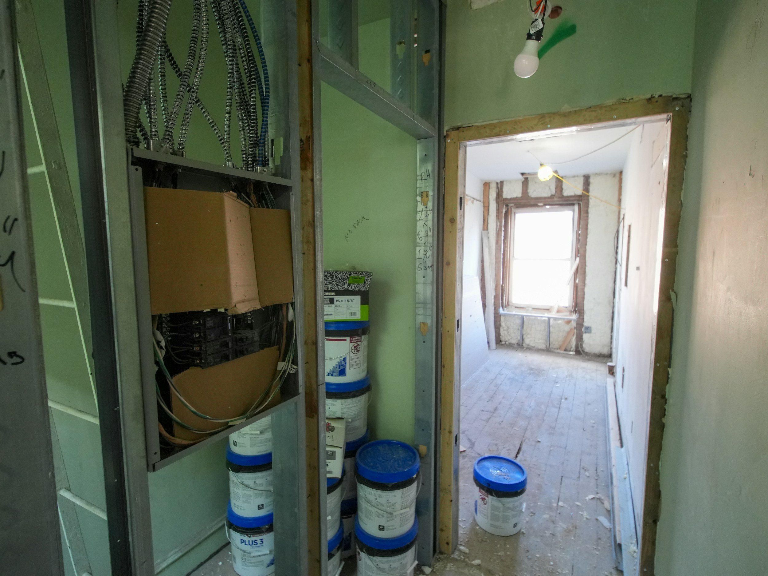 Interior of a building under construction showing electrical panel wiring, stacked paint buckets, and a doorway leading to a room with a window and construction materials.