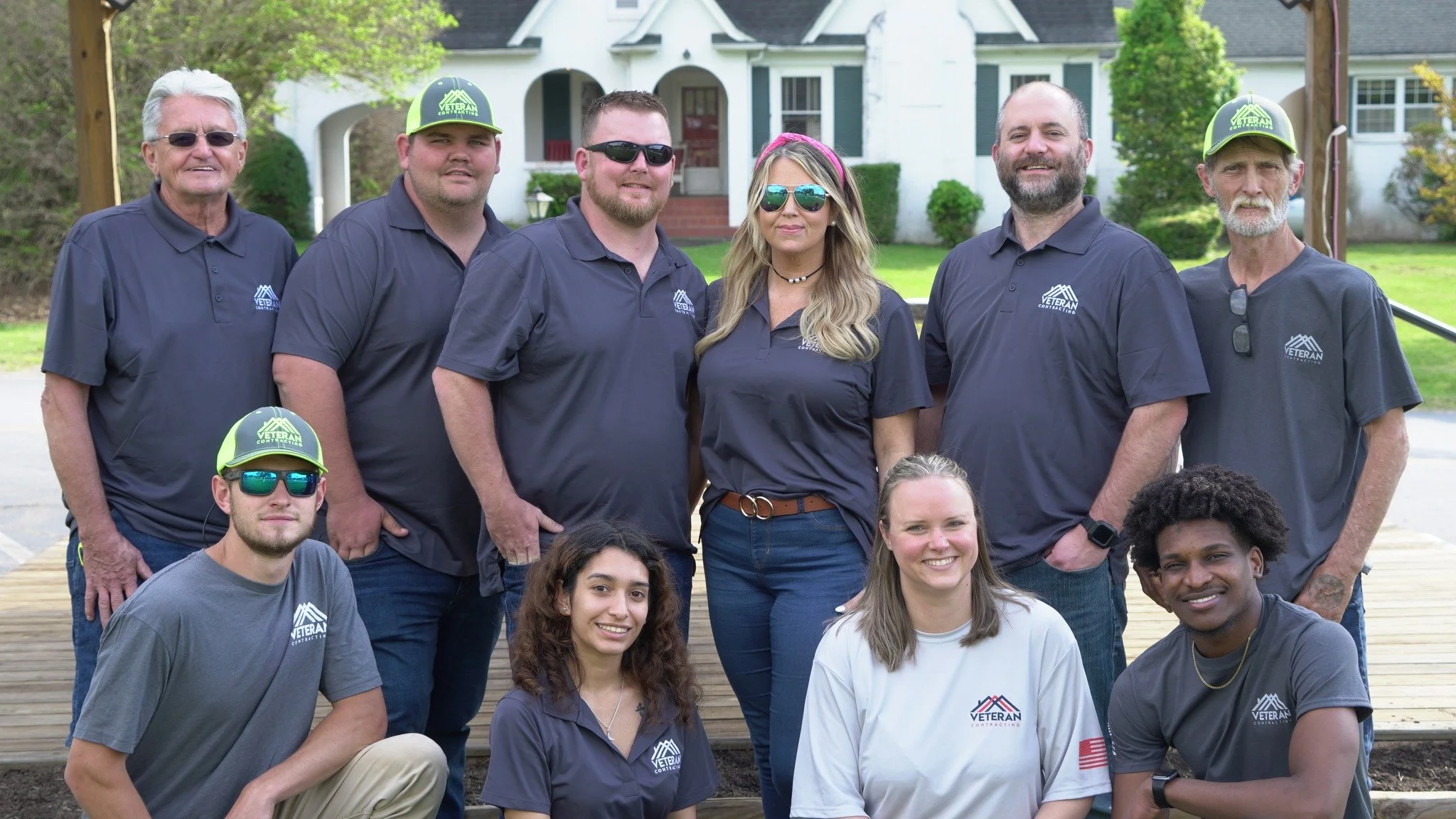 A group of eleven people, mostly wearing dark blue shirts with a 'Veteran Contracting' logo, standing outdoors on a wooden platform in front of a house, smiling at the camera.