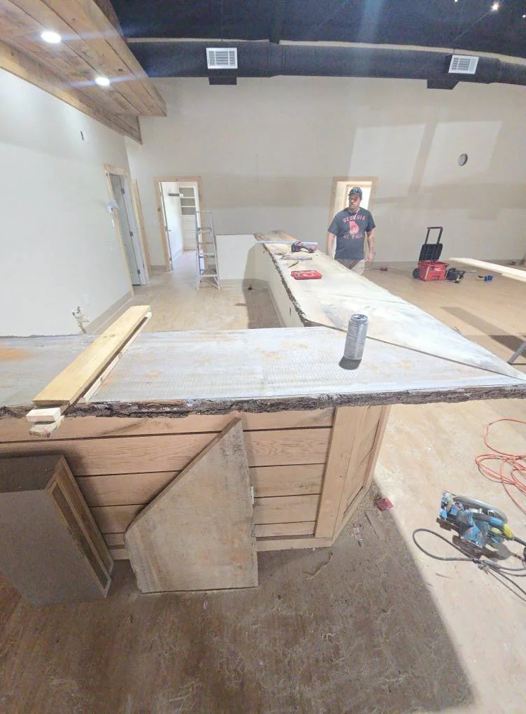 Interior of a room under construction with a worker standing near a partially built counter or island, construction tools and materials scattered around.