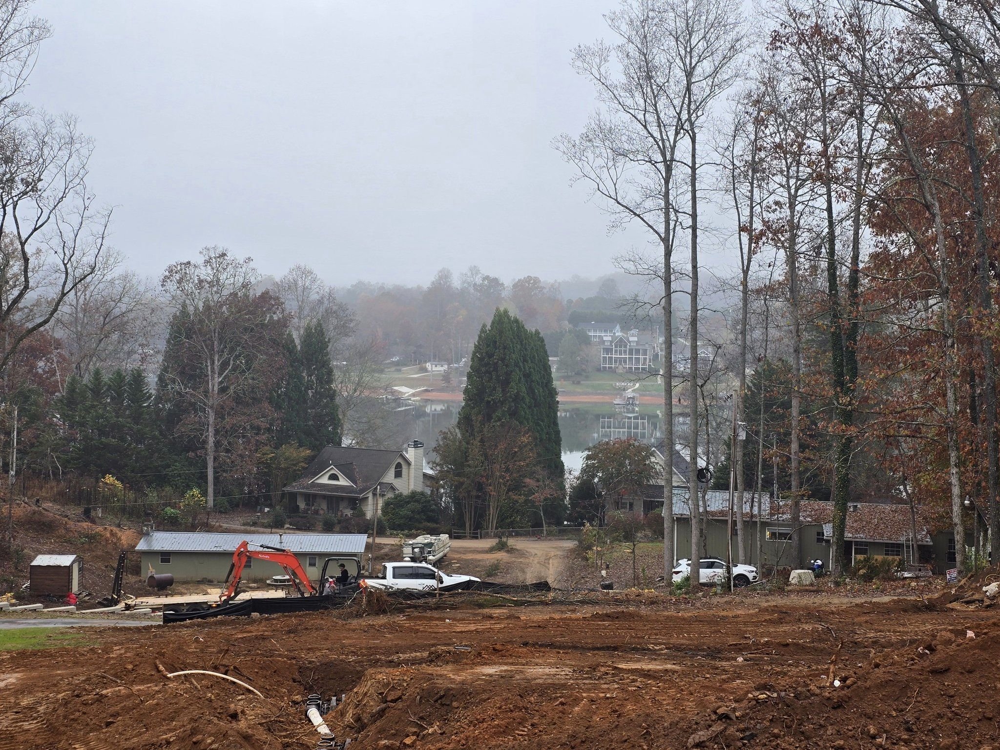 View of a construction site with dirt and an orange excavator in the foreground, residential houses, and a body of water in the background surrounded by trees and fog.