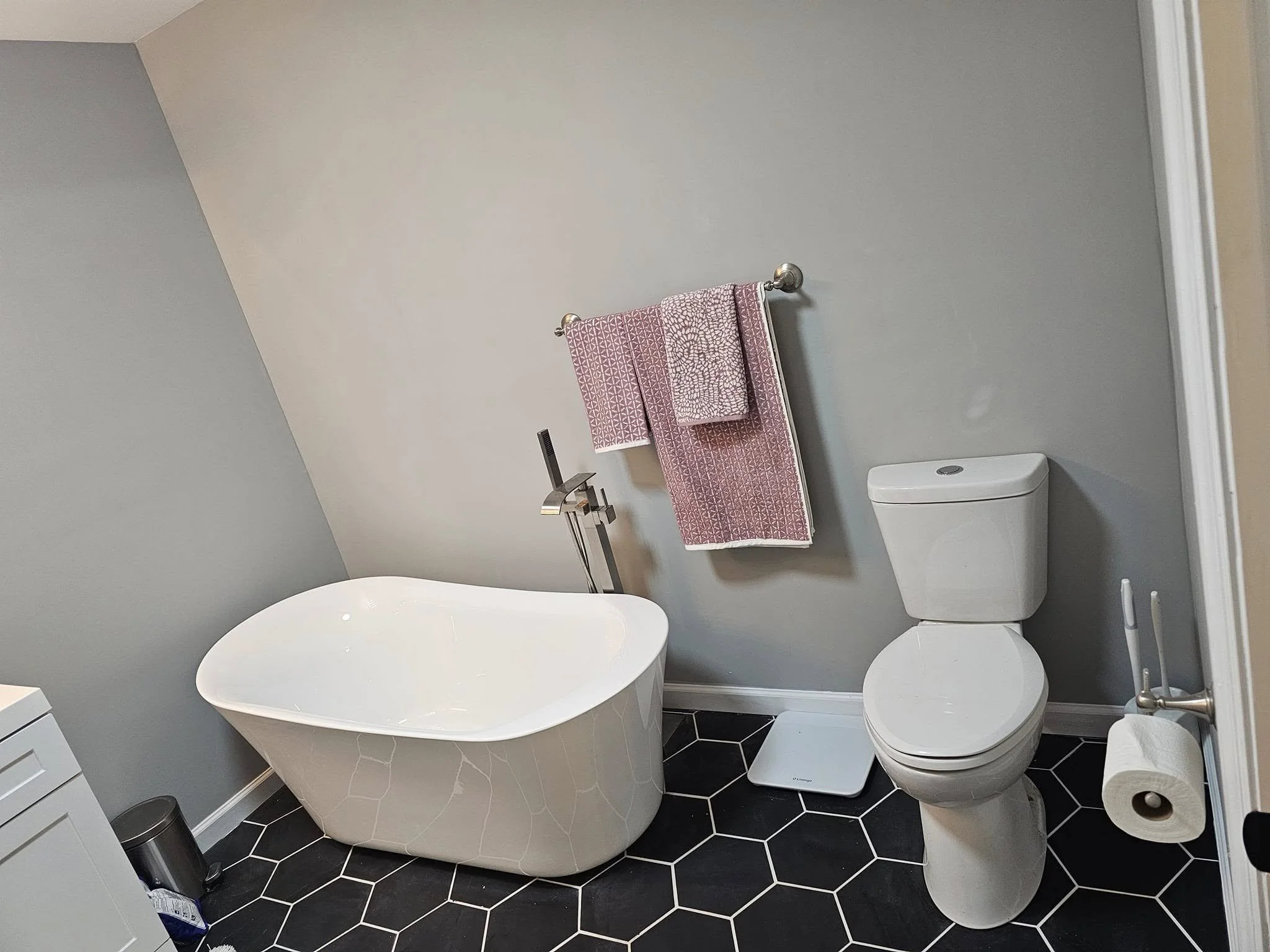 Bathroom with gray walls, black hexagon floor tiles, a white bathtub, a white toilet, a pink patterned towel hanging on a silver towel rack, a digital scale on the floor, and a toilet paper roll on a holder.