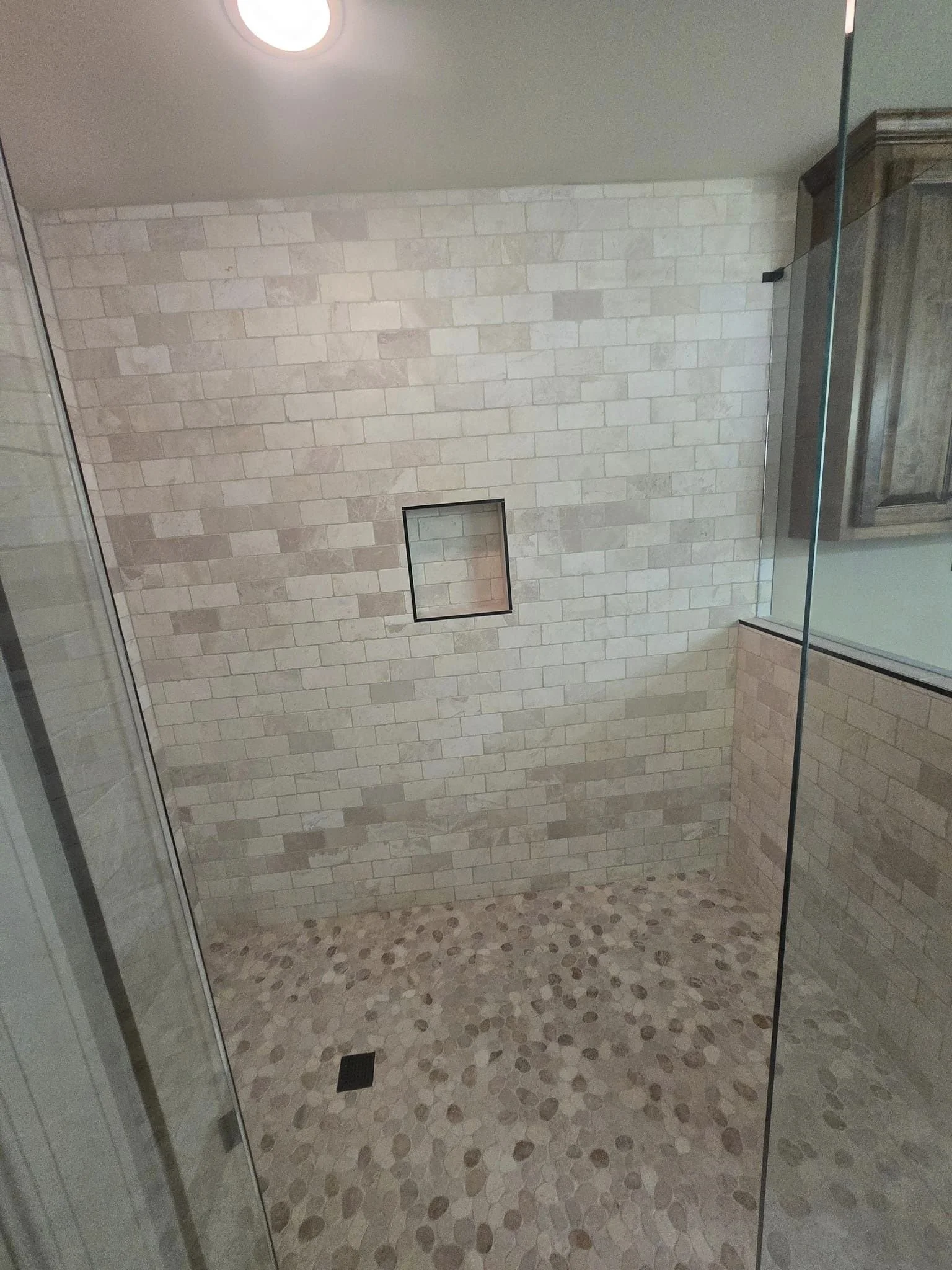 Shower with beige brick wall, pebble floor, small built-in niche, and glass door.