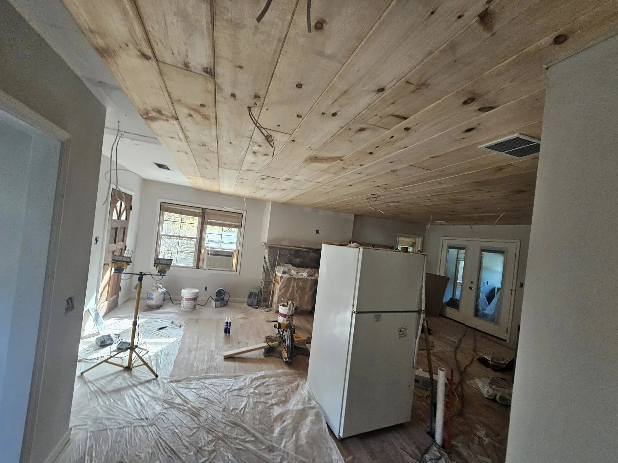 A room under renovation with wooden ceiling panels, construction tools, a refrigerator, window, and plastic sheeting covering the floor.