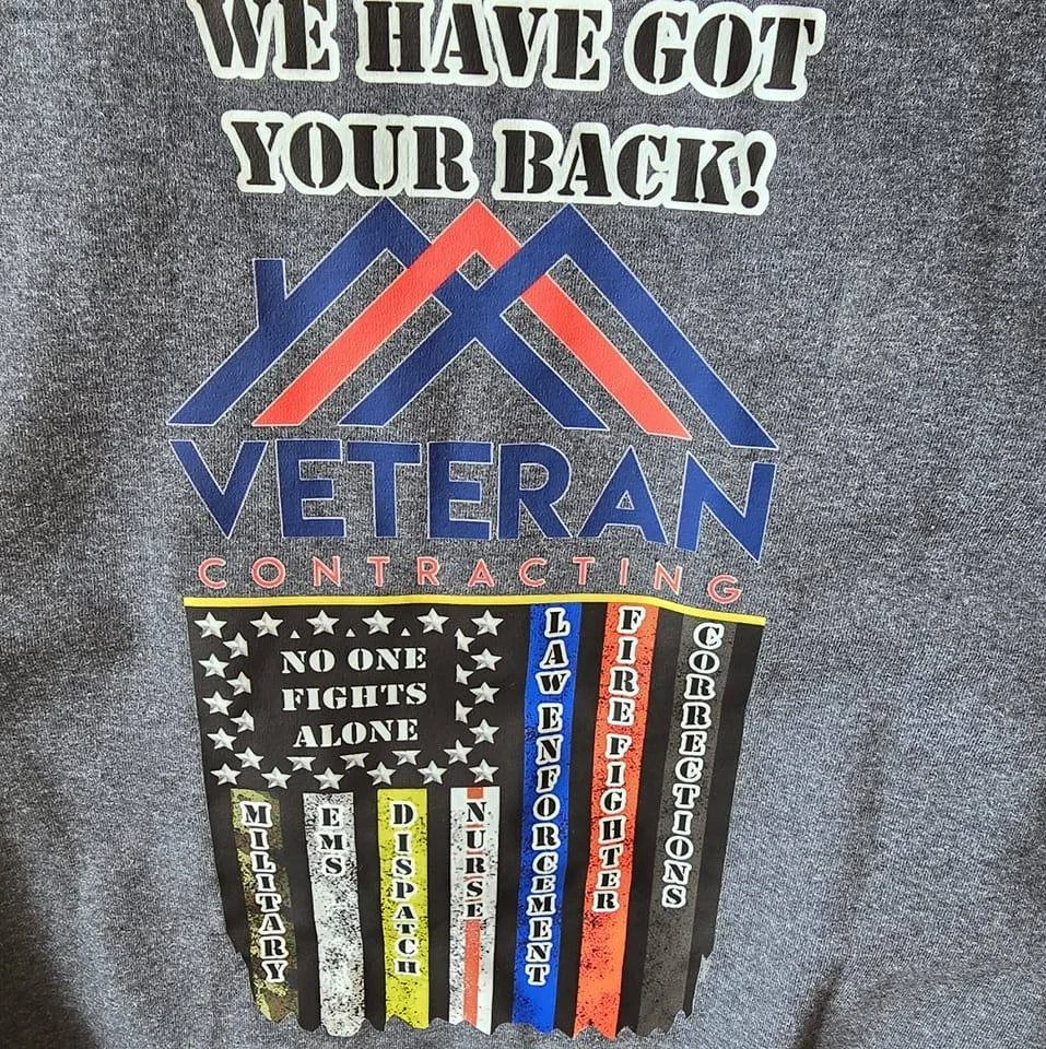T-shirt with patriotic-themed design and logo for veteran contracting. Text includes 'We Have Got Your Back!' and a graphic of a house roof. Below are symbols and words related to military and veteran services, such as 'No One Fights Alone,' 'Discharge,' 'Nurse,' 'Discharge,' 'Law Enforcement,' 'Firefighter,' and 'Corrections'.