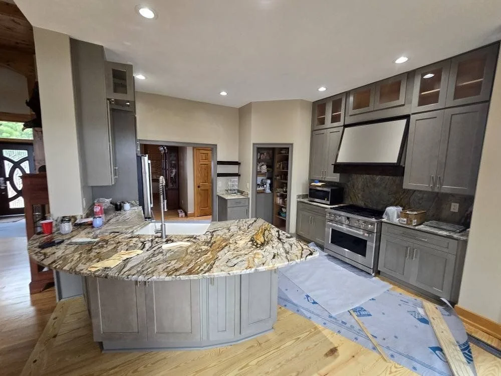 Modern kitchen with gray cabinetry, granite countertops, and a center island. Stainless steel appliances, including a stove and microwave, are visible. The kitchen appears to be under renovation or construction.