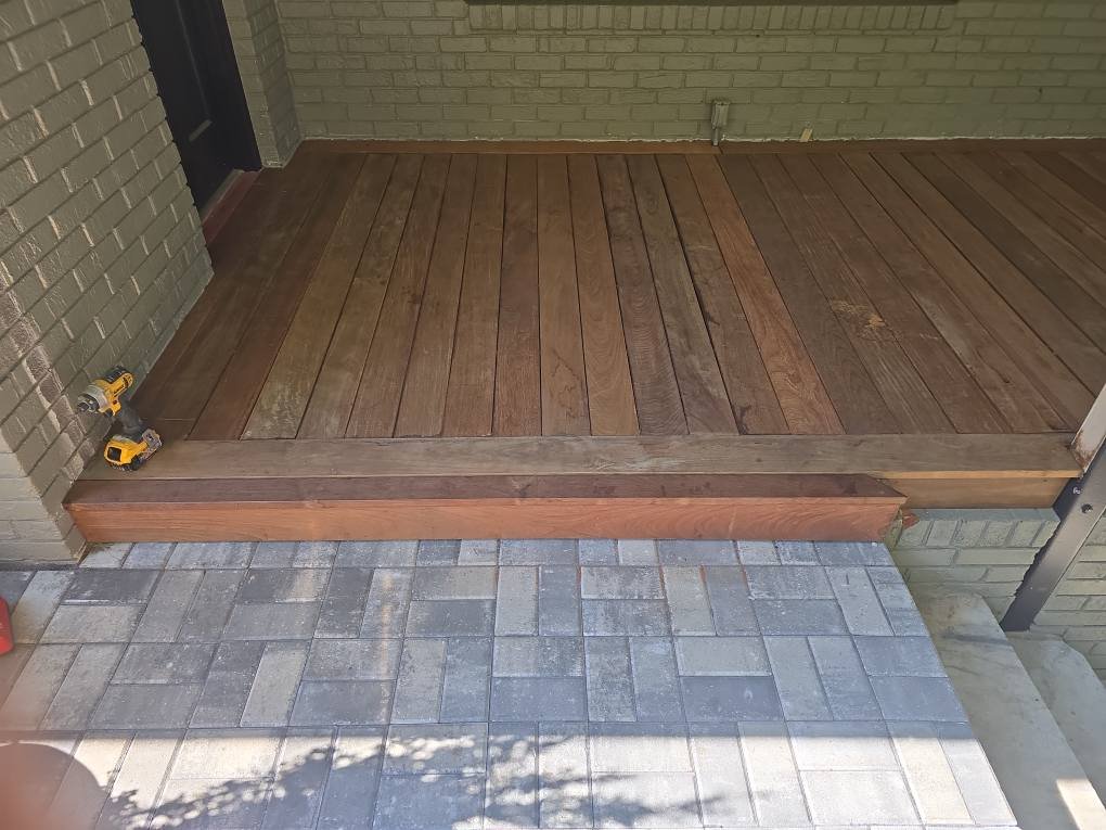 Wooden porch with brick walls and tools on the side, next to a paved walkway.