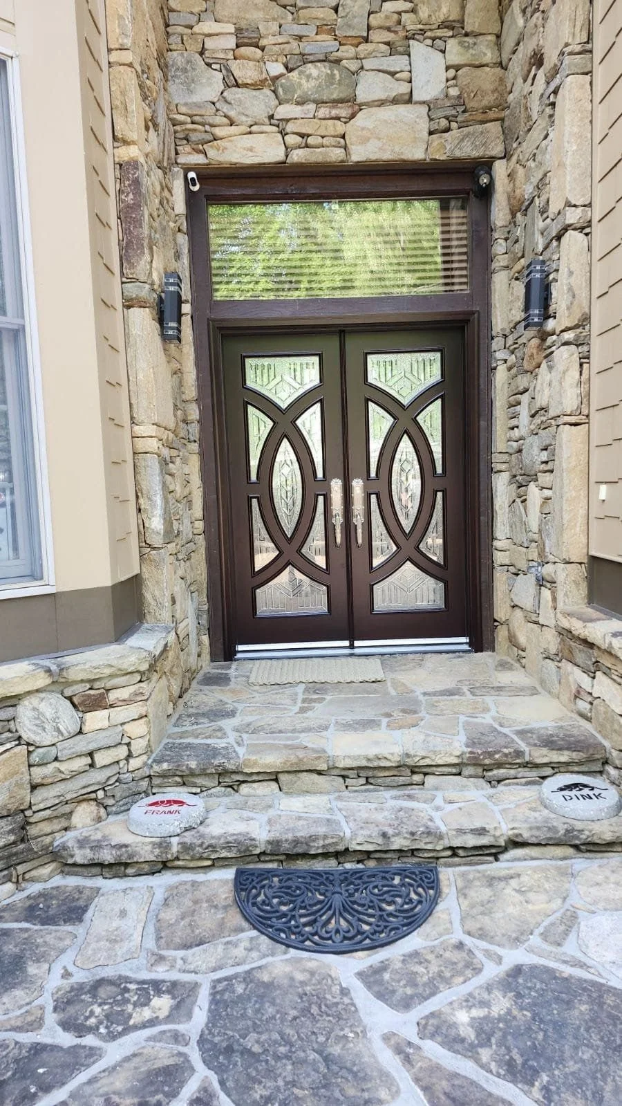 Front entrance with a decorative dark wood double door, stone steps, and stone wall framing around the door. There are two round stone markers with dog paw prints and names, a doormat, and a decorative black doormat on the stone pavement.