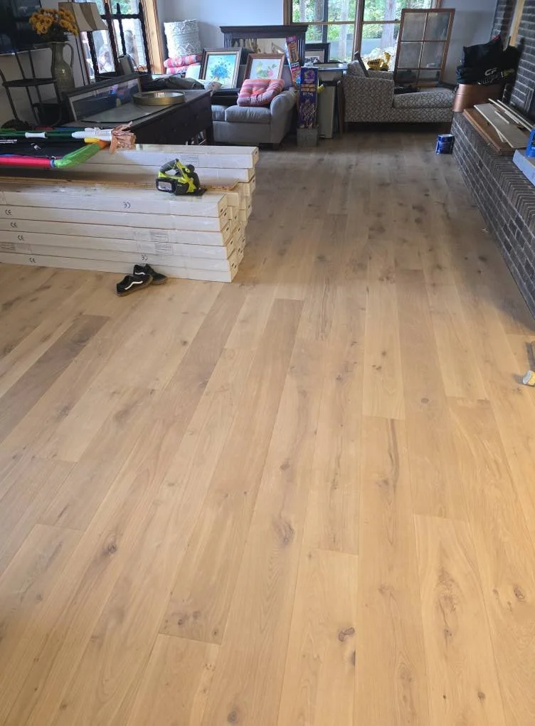 Undeveloped wooden flooring in a living room with household items and furniture on the edges, including framed pictures, a sofa with pillows, and construction tools on a pile of wood planks.