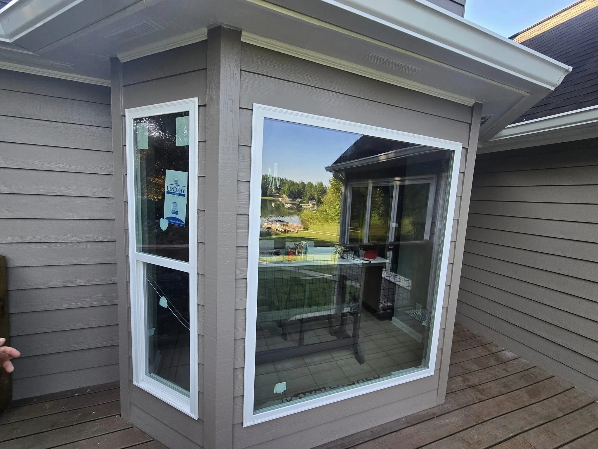 A small, enclosed porch or sunroom with large windows, attached to a house on a wooden deck. The house has beige horizontal siding and a white roof trim. Through the windows, a lake with trees and some outdoor furniture is visible.