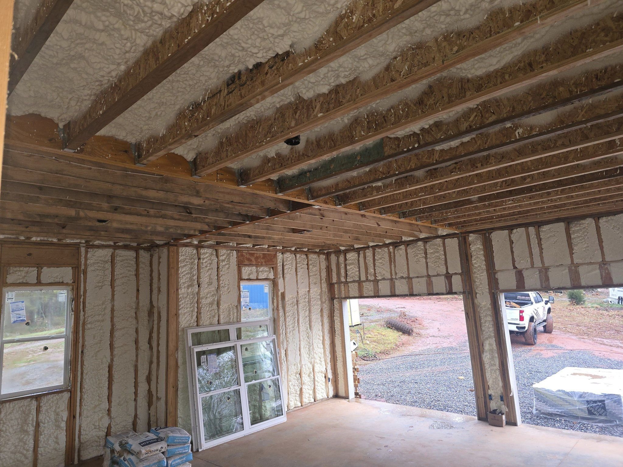 Interior of a house under construction, with exposed roof trusses, spray foam insulation, and unfinished walls. Construction materials and windows are visible.