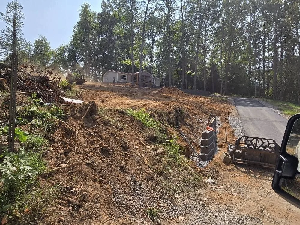 A house situated on a hilltop with trees in the background, next to a road that is under construction with gravel and blocks.