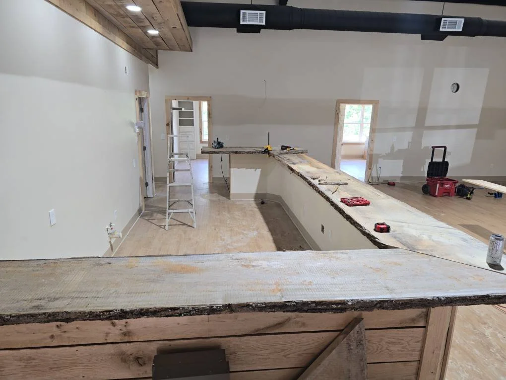 Interior of a house under construction, showing a partially finished kitchen island with tools and construction materials on it, open doorways, and windows with natural light.