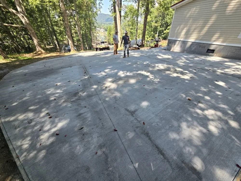 Construction workers working on a concrete foundation outside near a house, with trees and a distant mountain in the background.