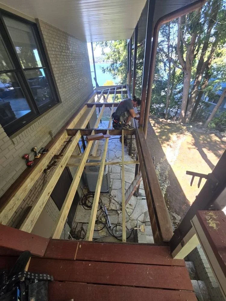 A man working on a wooden balcony construction, with tools and wood planks, overlooking a wooded area and water. The view is from an upper floor of a building.
