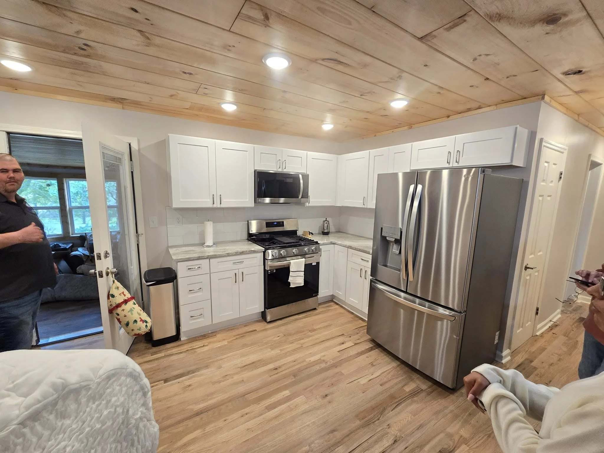 Modern kitchen with white cabinets, stainless steel appliances, and wooden flooring, with people partially visible on either side.